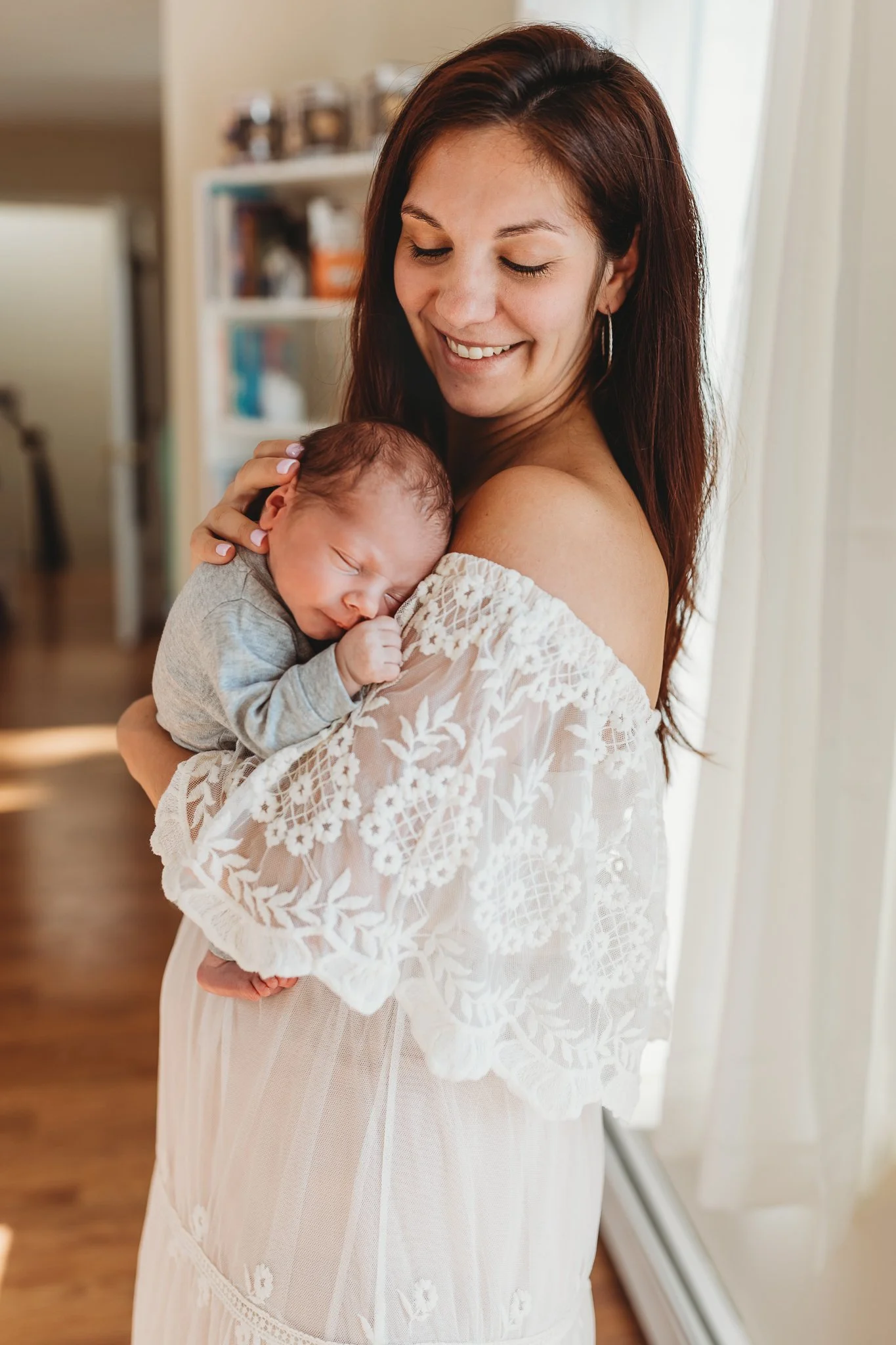 mom in boho ivory lace dress who is holding baby boy in grey onesie and he is sleeping she is by a window and is smiling