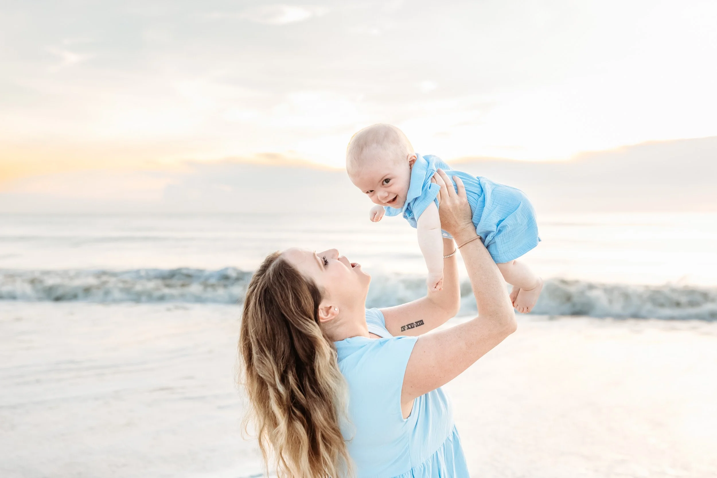 mother lifting baby boy in the air who is laughing and the ocean and sunrise is behind them