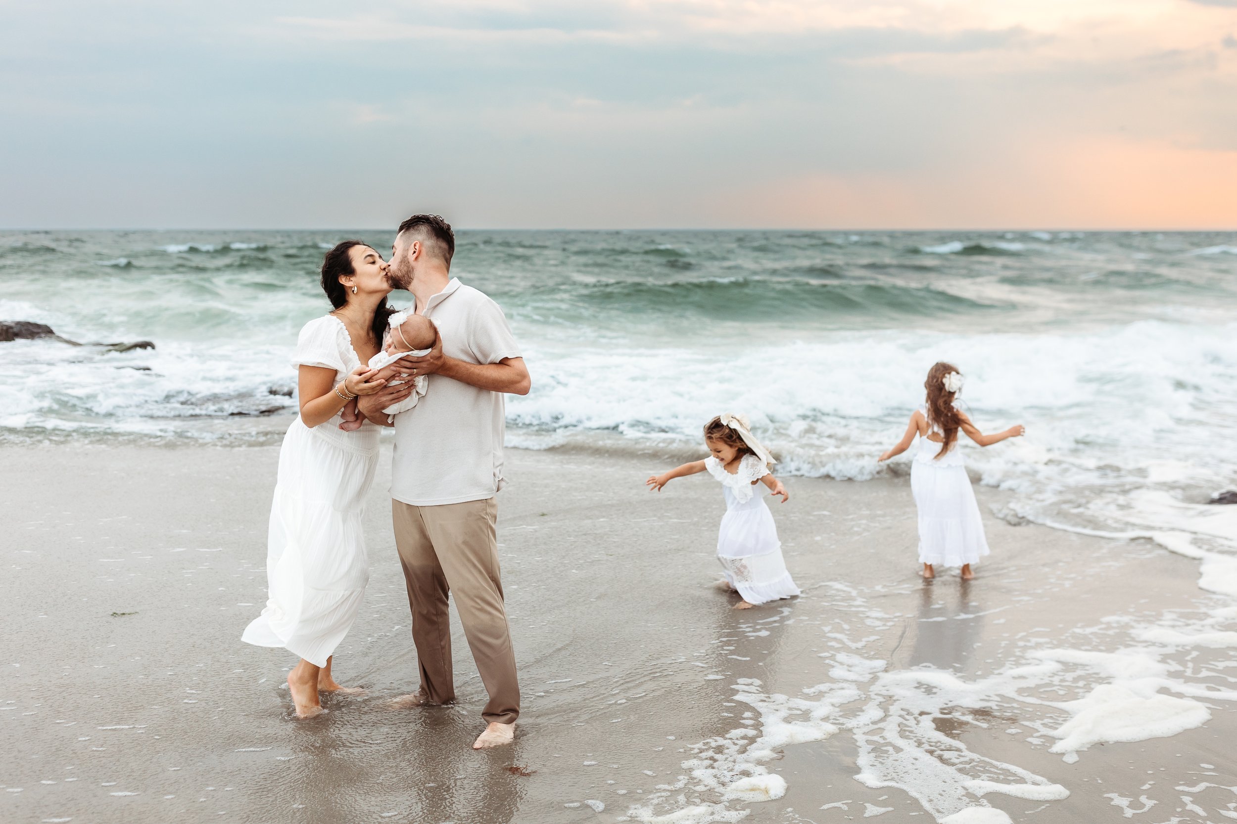 husband and wife kissing nn the beach while holding their newborn and their toddler girls are playing in the ocean