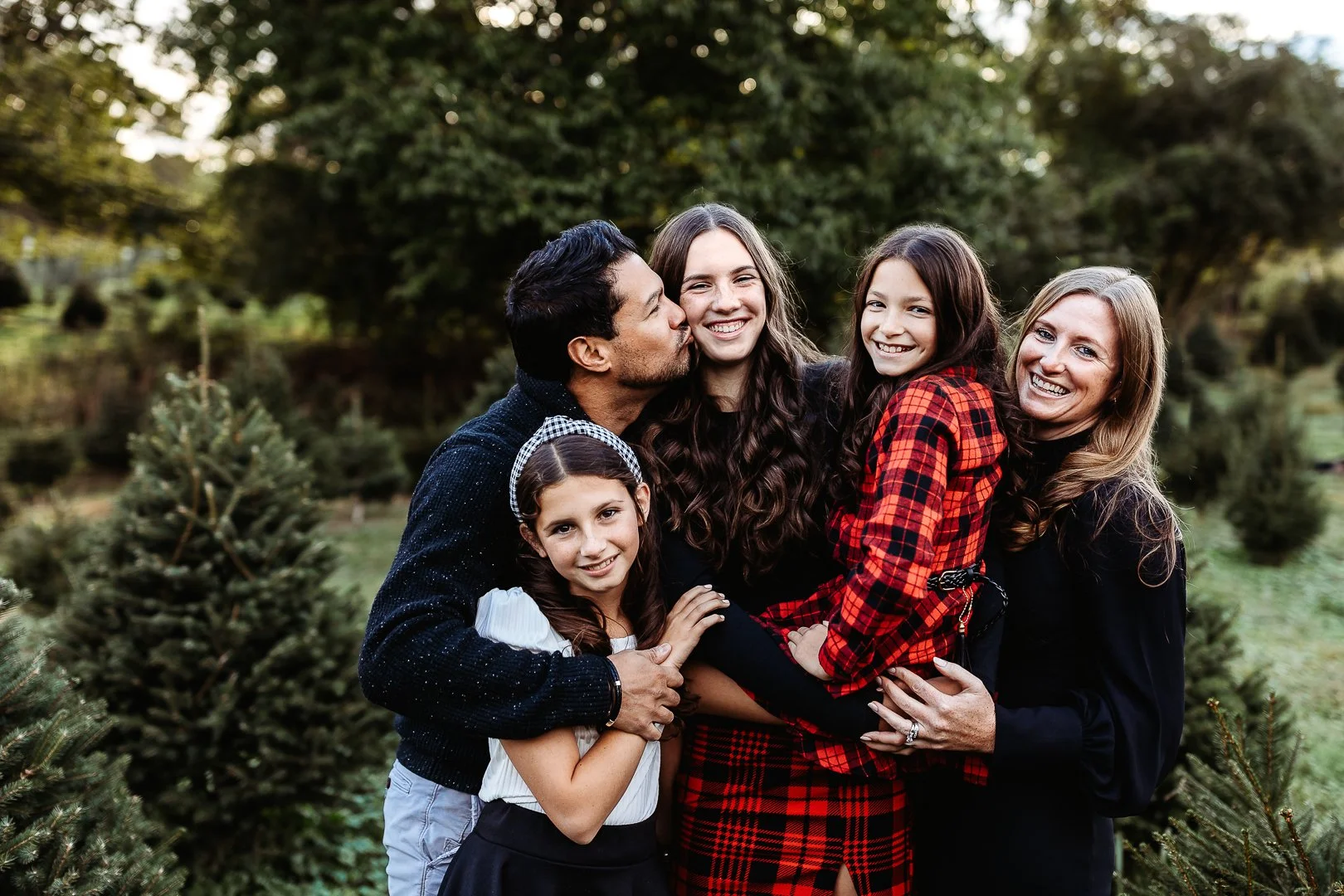 family in red and blaick buffalo plaid embraced and laughing at a tree farm