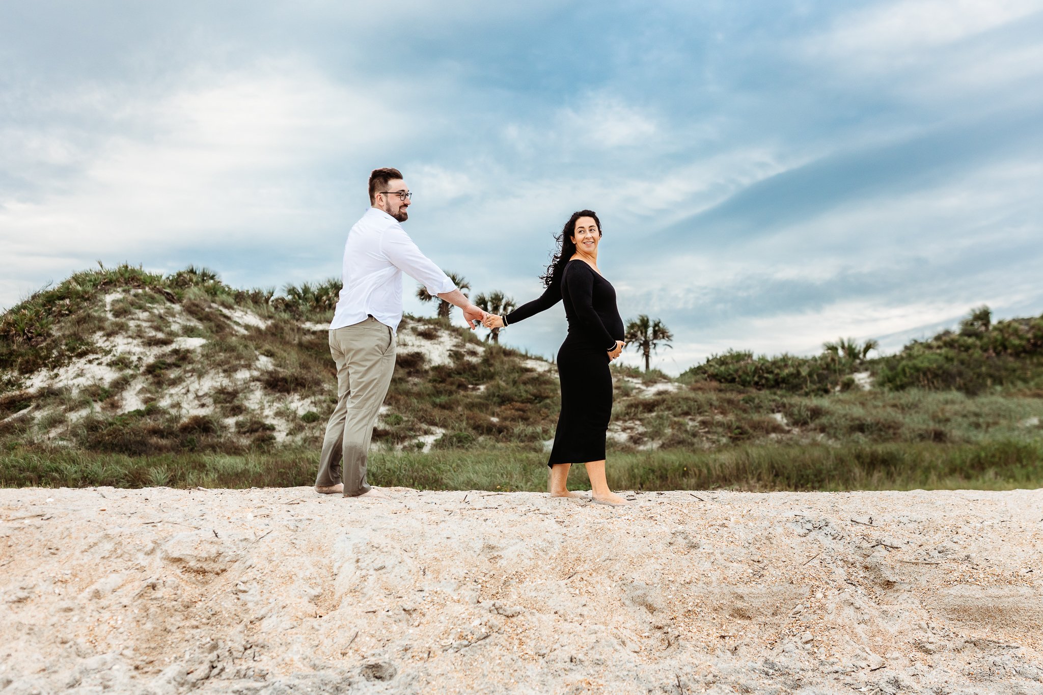 couple on the top of a dune walking single file with blue sky and palms behind them and woman is holding pregnant belly