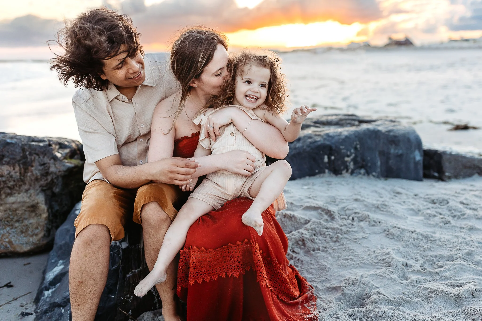 family embraced while sitting on a jetty at Villano beach and holding and kissing their toddler for a sunset family photo session