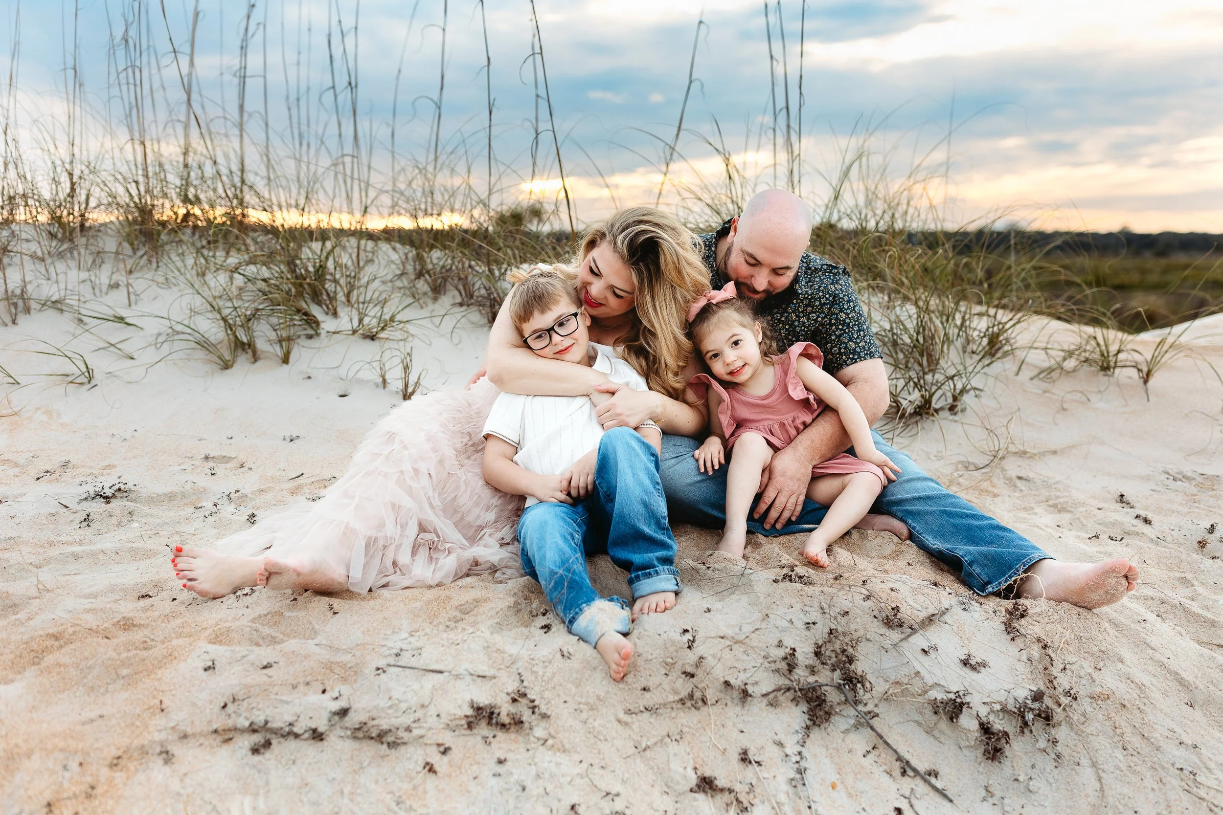 family seated on a dune at the beach with toddler children on their lap and they are kissing them at sunset