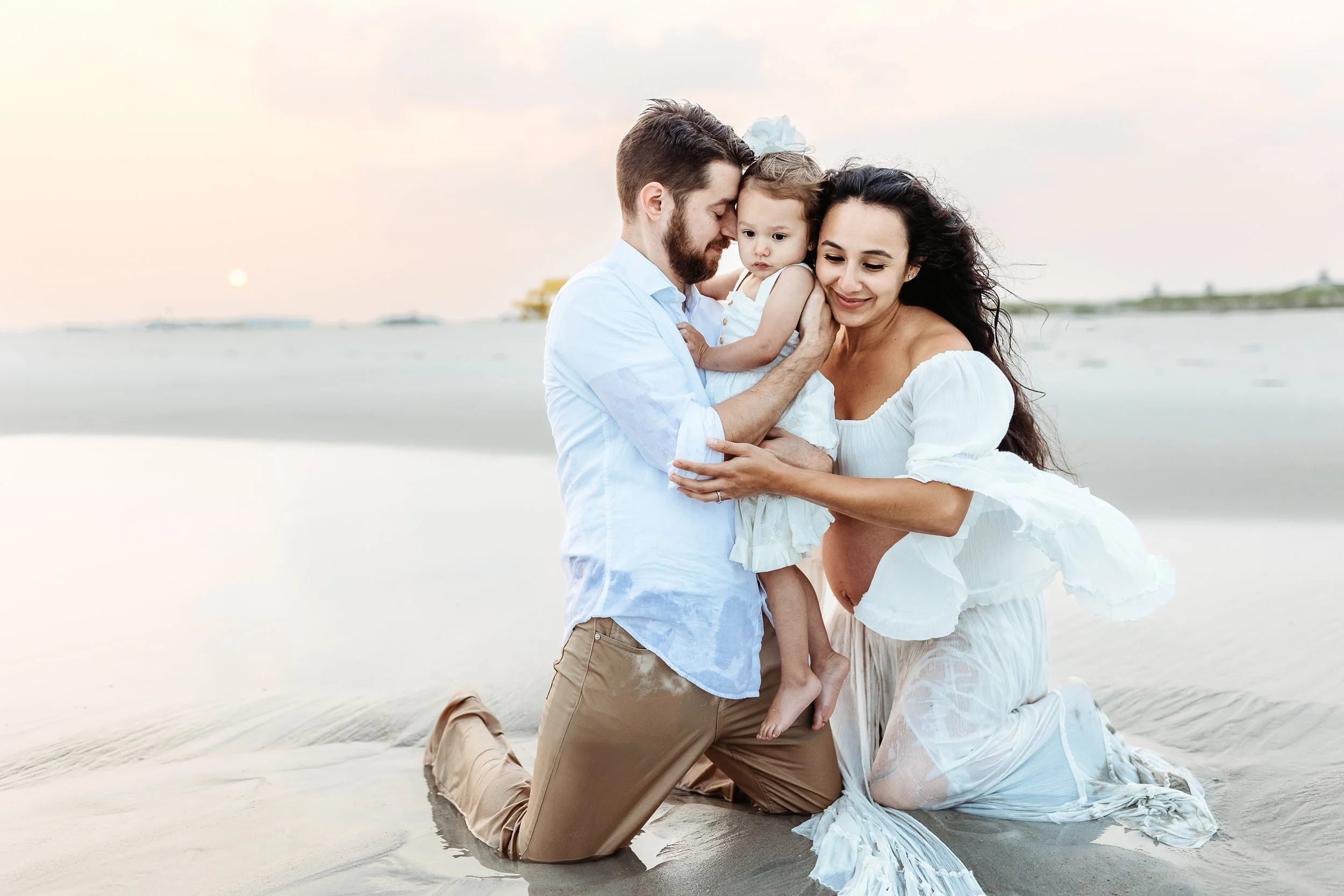 mother and father kneeling in the ocean and embracing toddler daughter mom is pregnant and belly is exposed