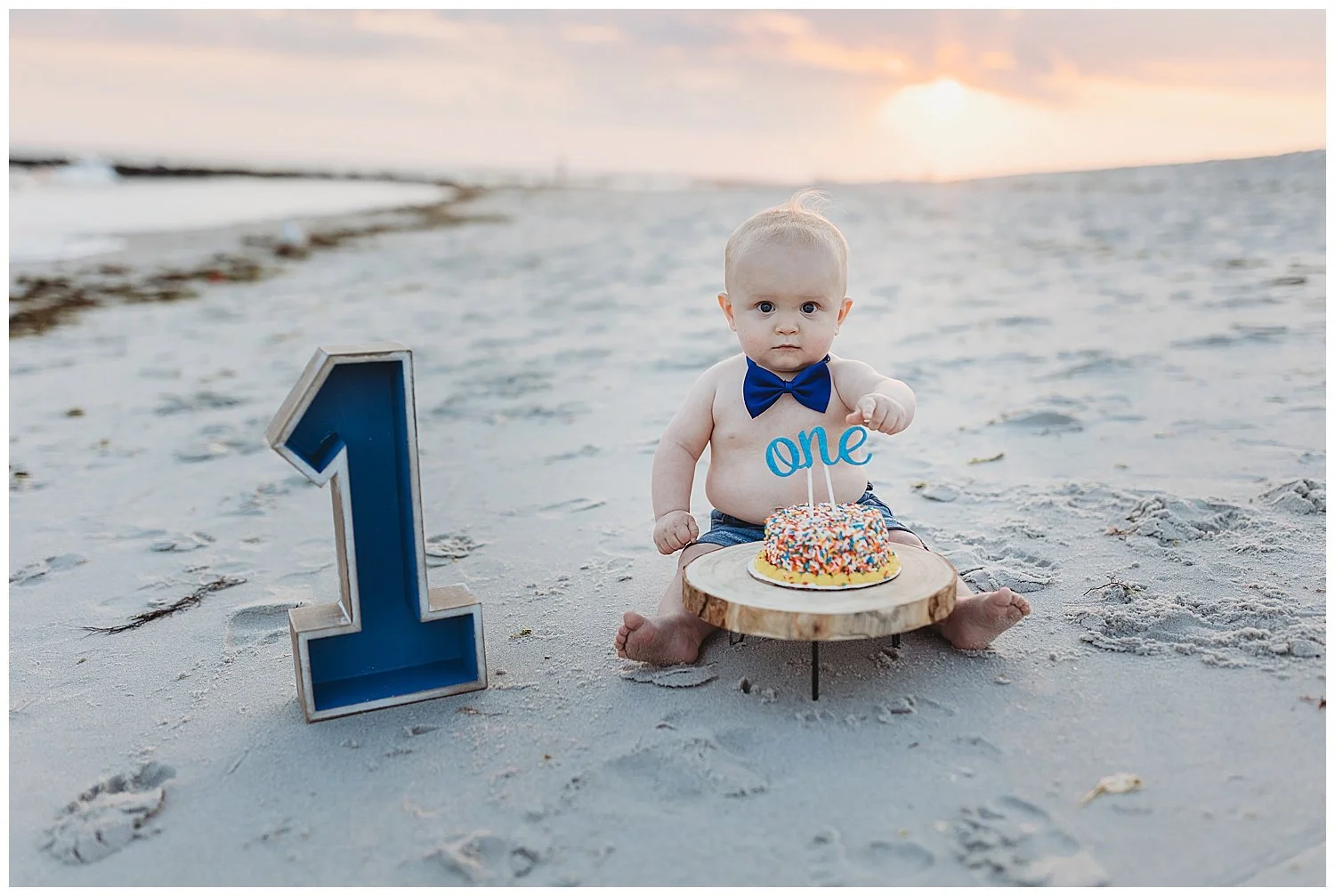 Baby boy with bow tie on the beach with no shirt on and a cake in front of him