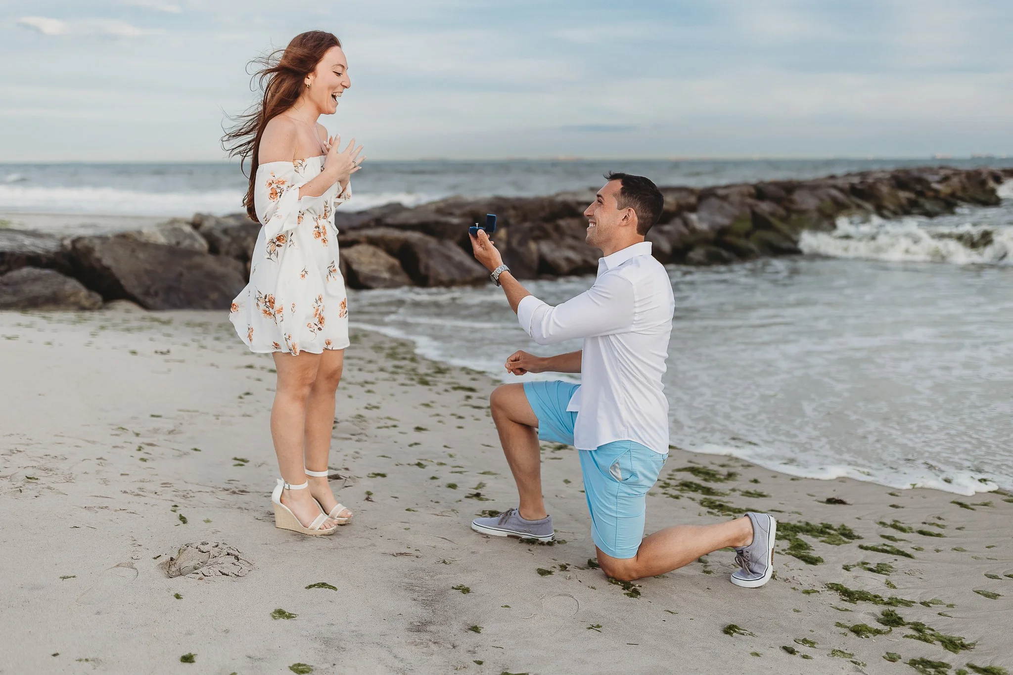 man on knee proposing to a girl in front of a jetty and she is shocked