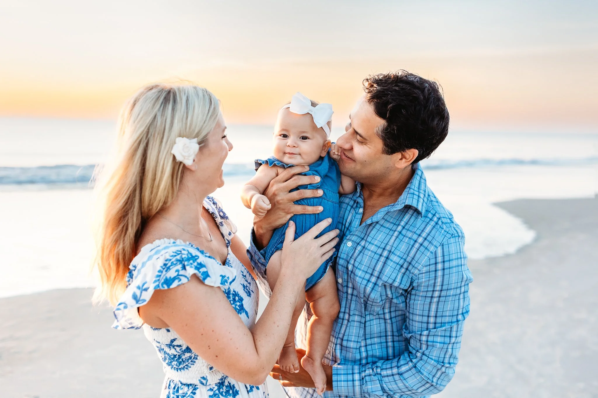 dad holding baby girl and mom is looking at her the baby is framed between the two of them and the sun is rising over the ocean