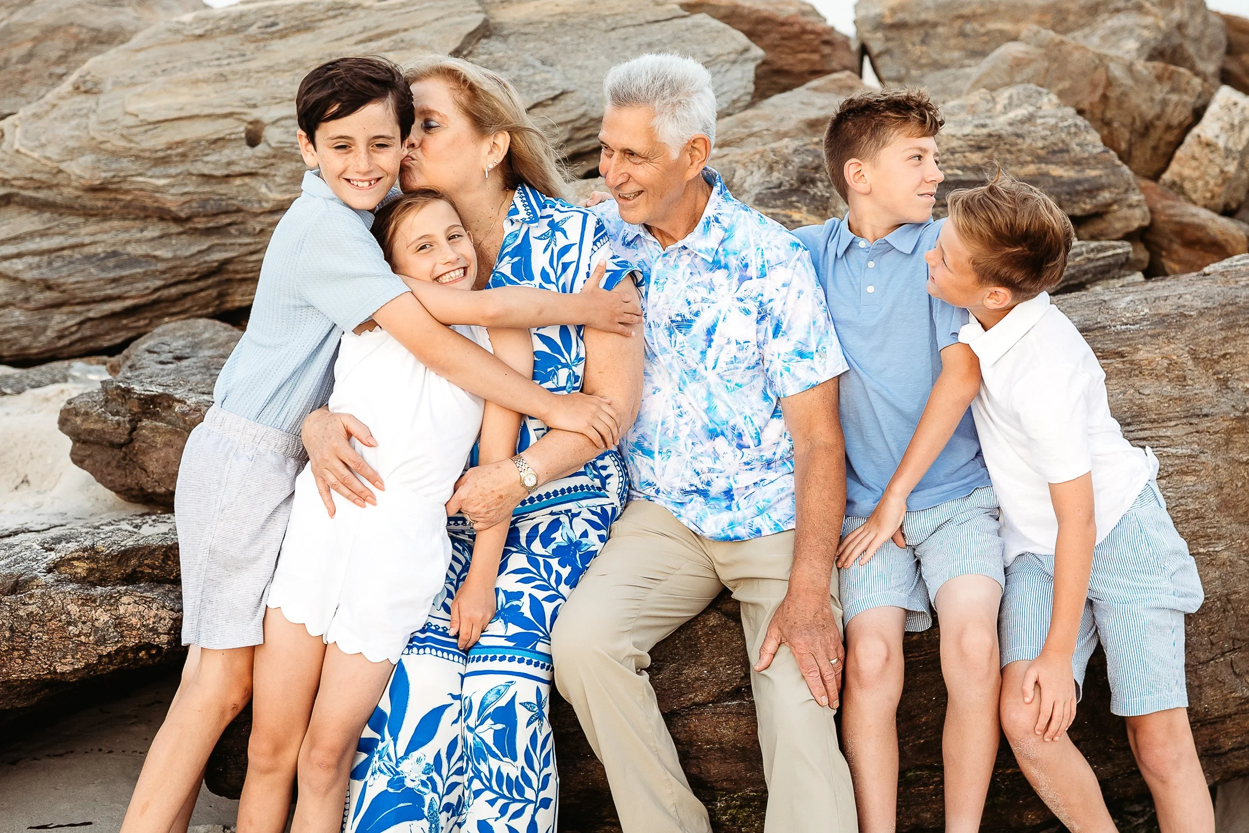 grandparents seated on a jetty and grandma is kissing one grandson while grandpa watches.  The other kids are laughing.  