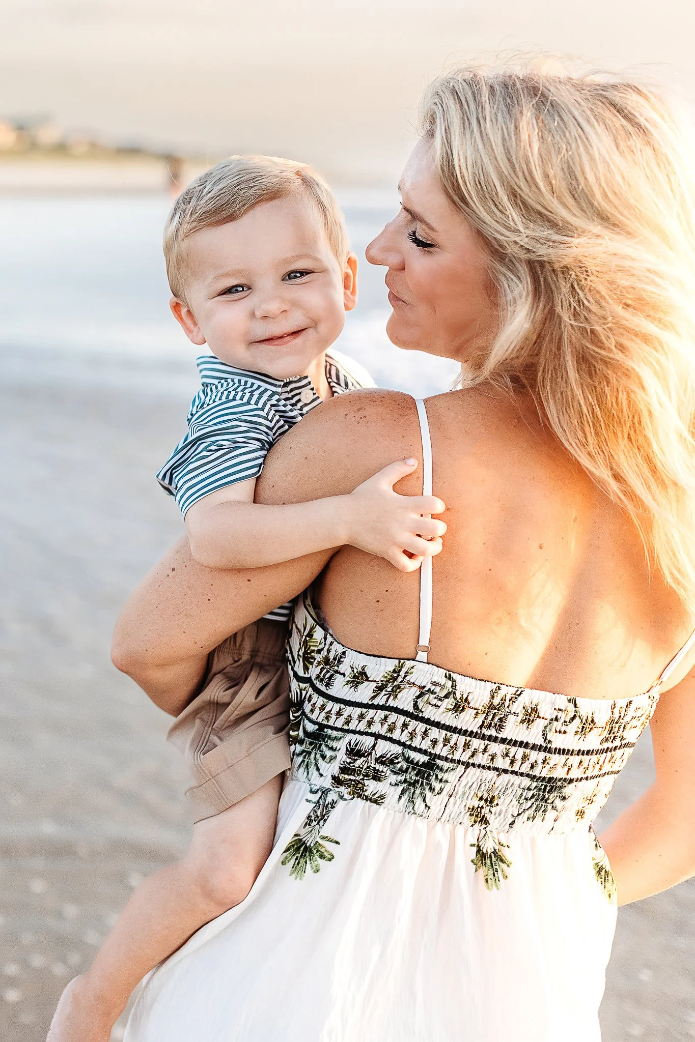 A woman with blonde hair holding a smiling young boy with blonde hair at the beach during sunset.