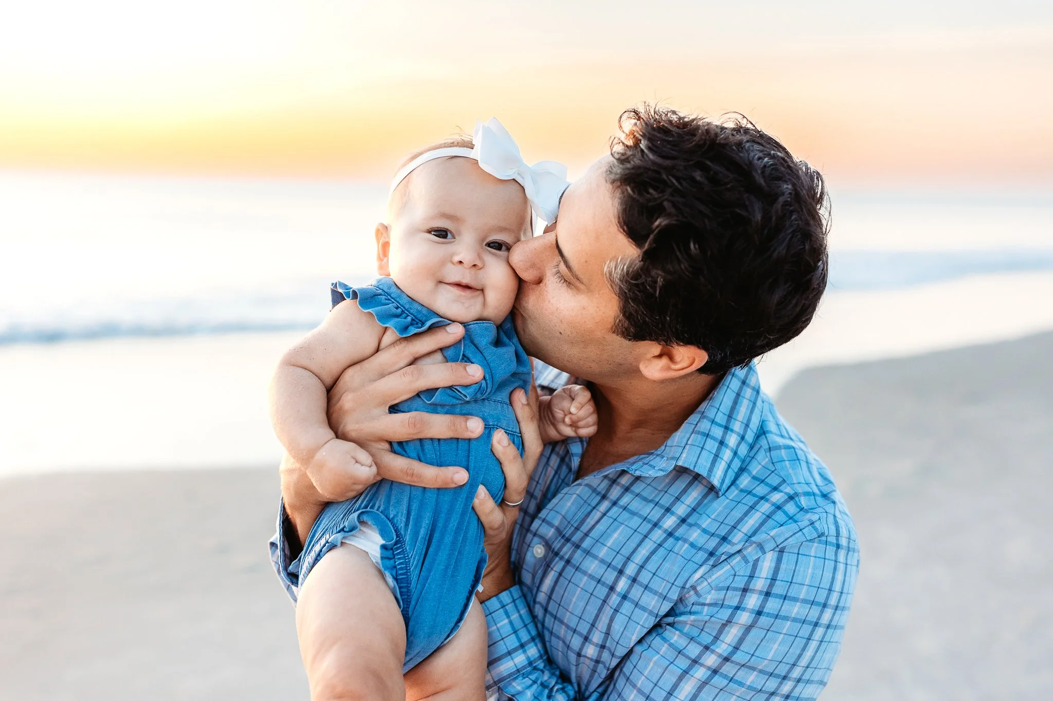 dad kissing his baby girl in a denim romper with the ocean at sunrise behind him