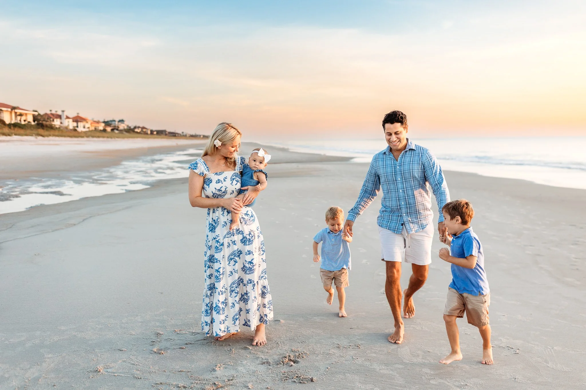 dad running on the beach in Jacksonville at sunrise with toddler boys while mom is in the foreground holding a baby girl and watching