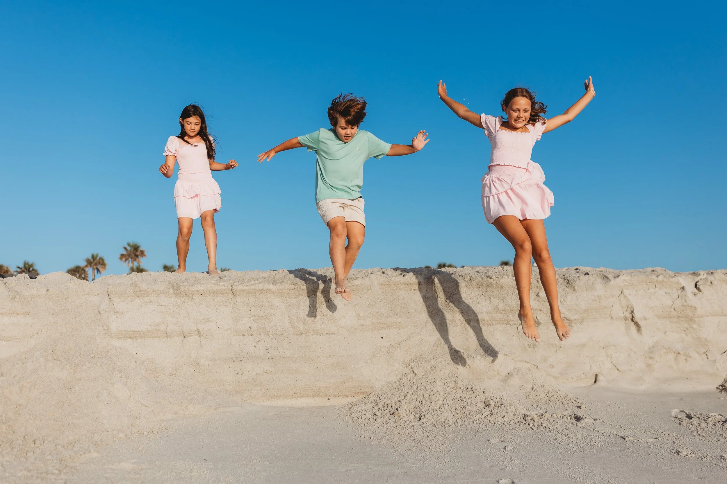tweens jumping of a sand hill and they are laughing and the sky is bright blue behind them