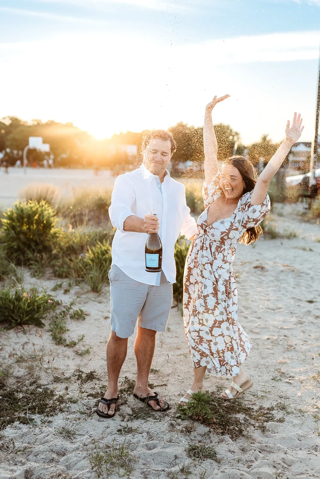 man popping bottle of champagne while girlfriend raises her hands and celebrates after they get engaged on the water in Jacksonville