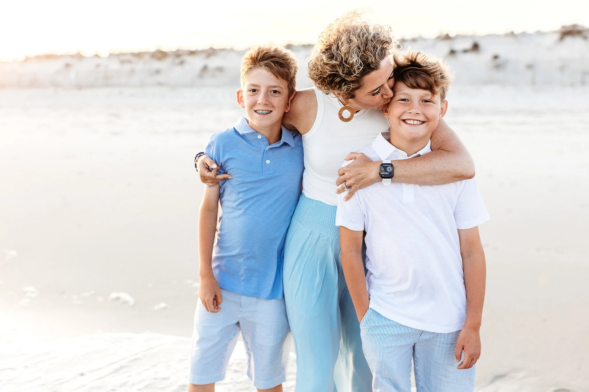 mother holding her boys and kissing them on the beach they are dressed in blue and white 