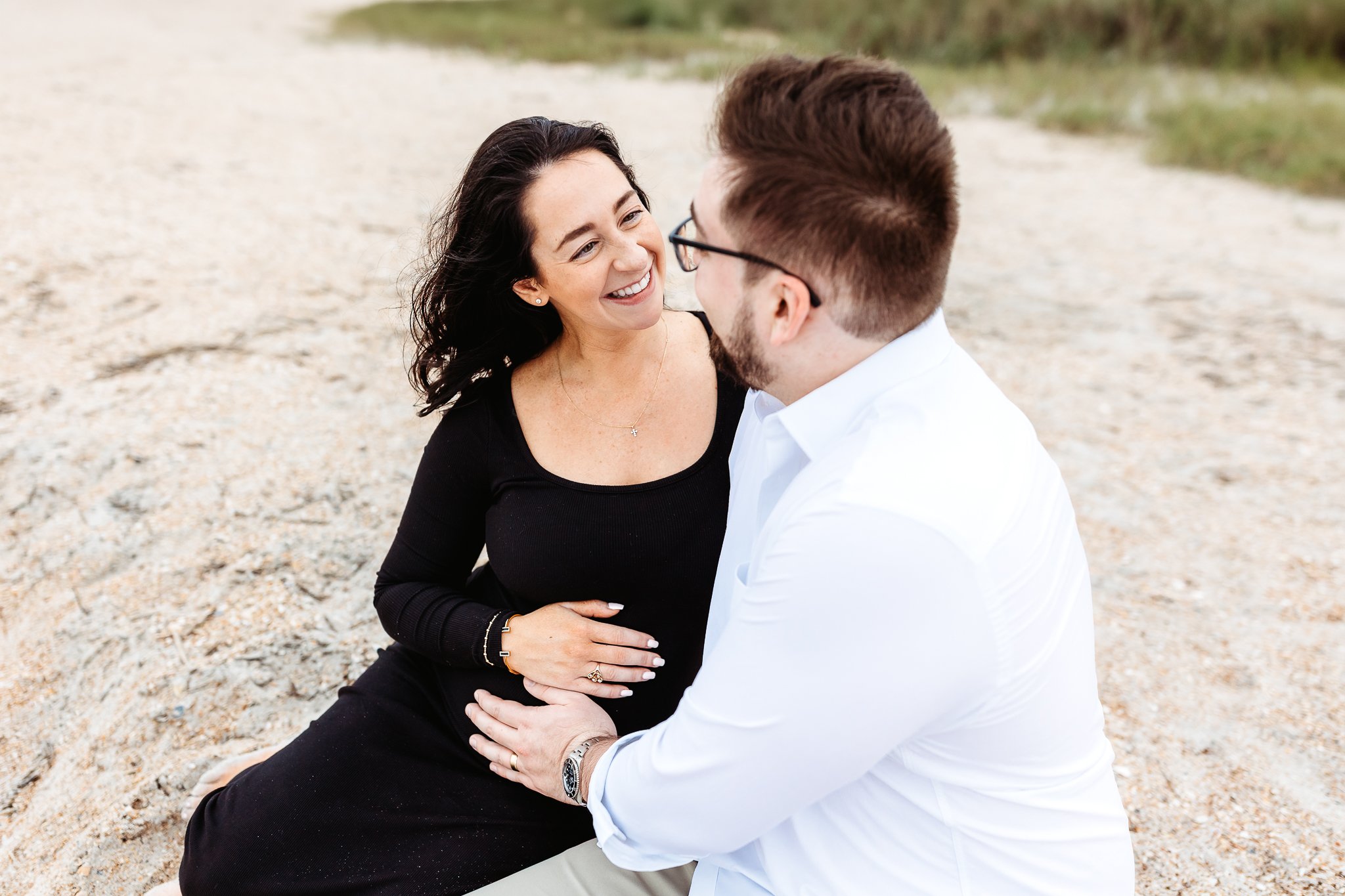couple looking at each other while seated not he dune and laughing and husband is hiding expecting mom's belly