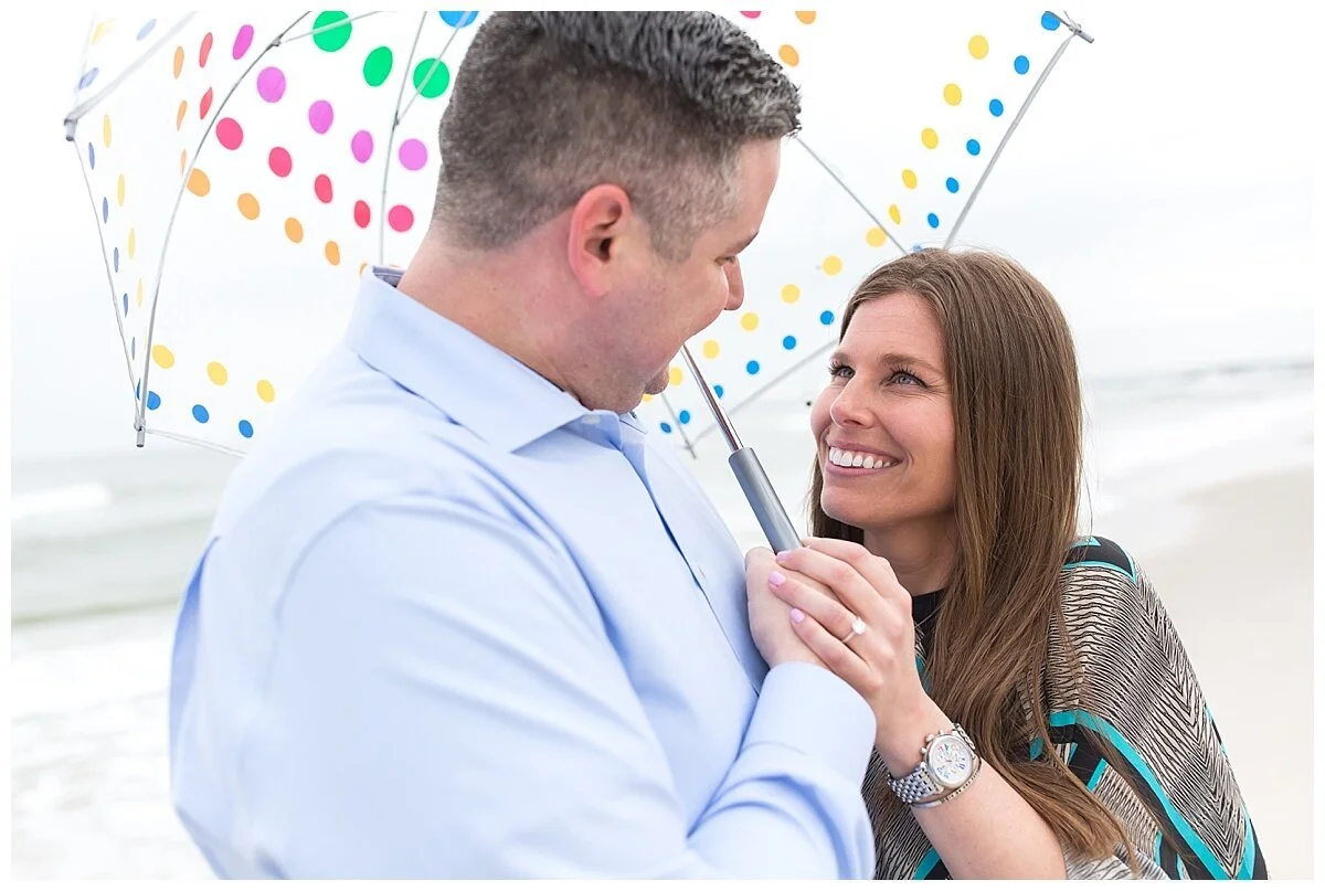 newly engaged couple under a polka dot clear totes umbrella looking at each other and they are on the beach