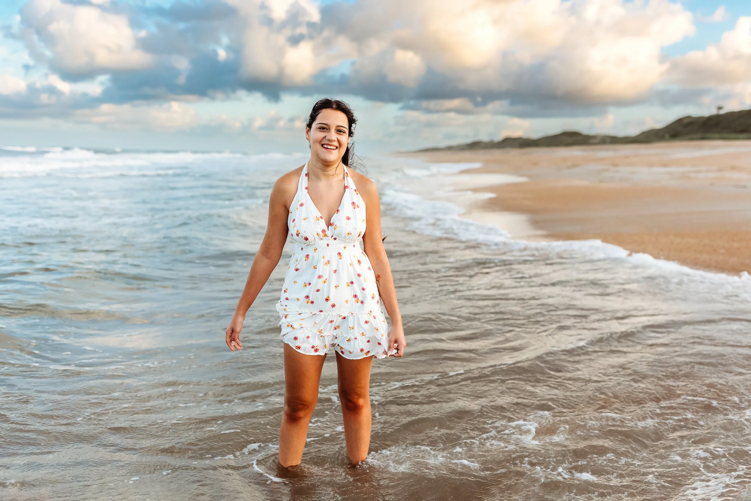 girl laughing in the ocean in floral dress with pastel sky behind her