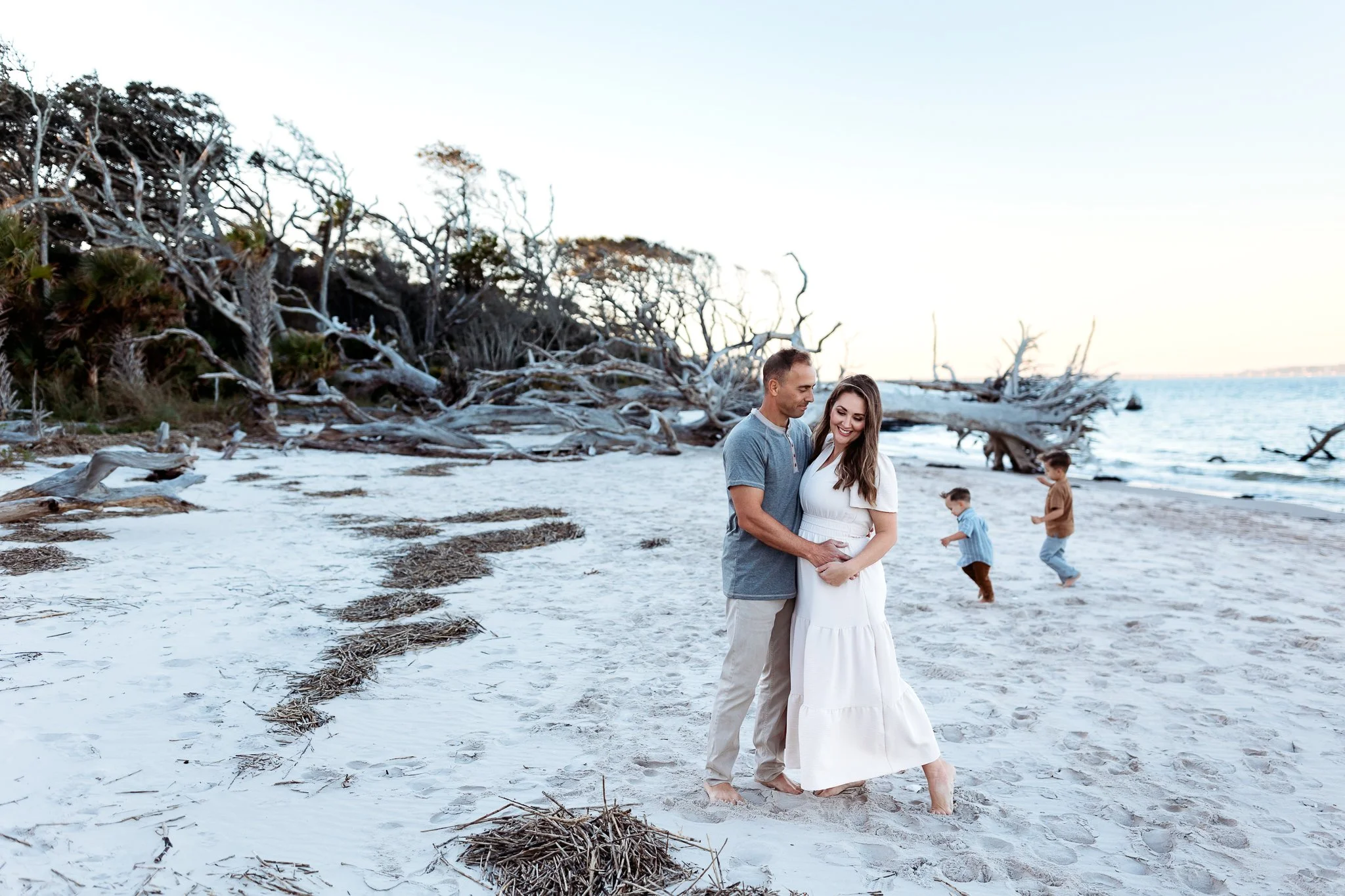 Parents embraced romantically on the beach while their toddler children are chasing each other on boneyard beach during a photo session