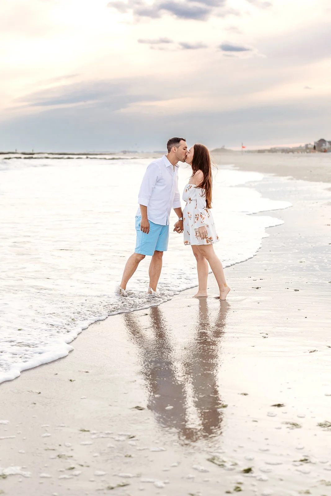 A couple sharing a kiss on the beach at sunset, holding hands, with their reflection visible in the wet sand.