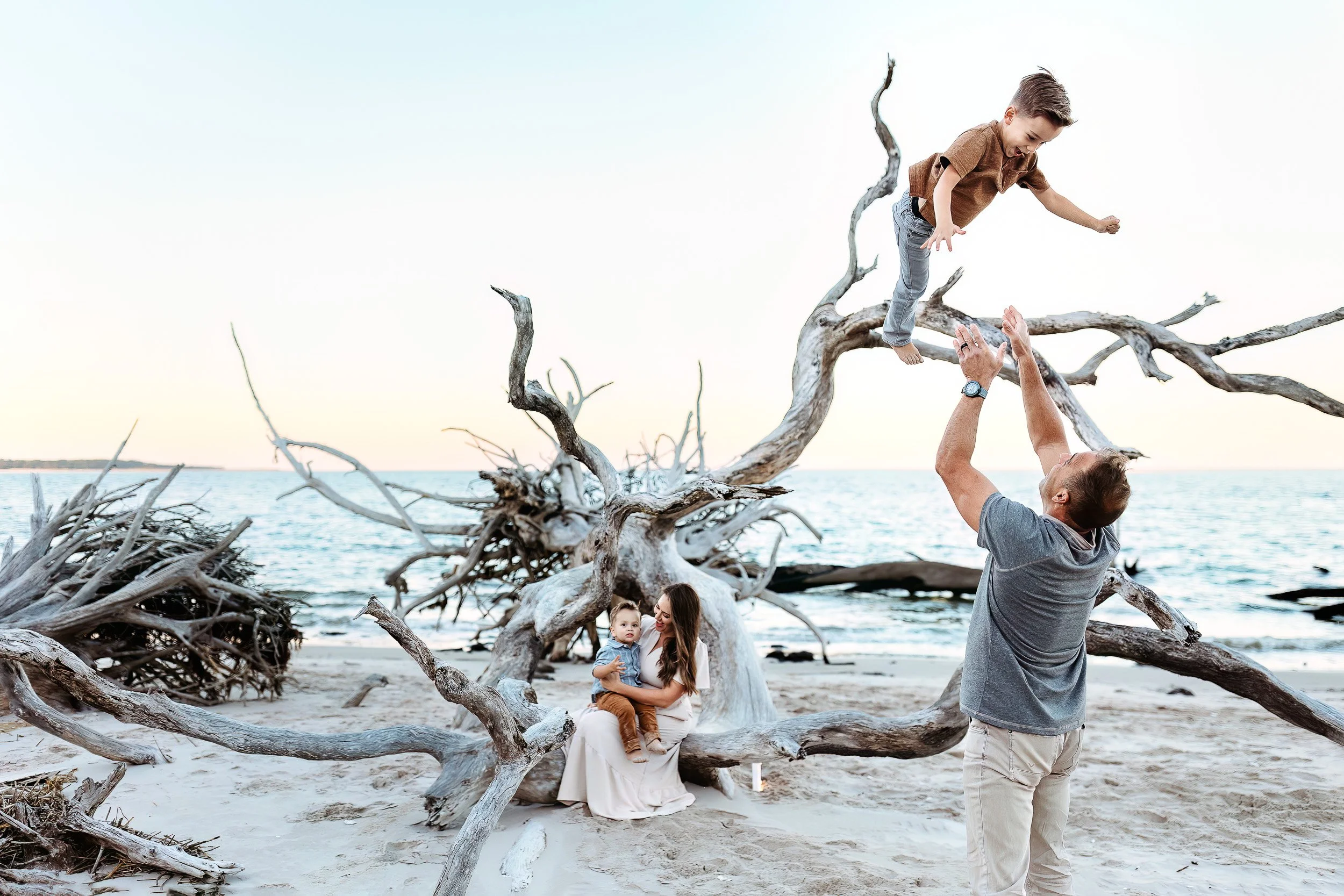 A family of four standing on a fallen large tree trunk on a beach during sunset, with leafless trees in the background.