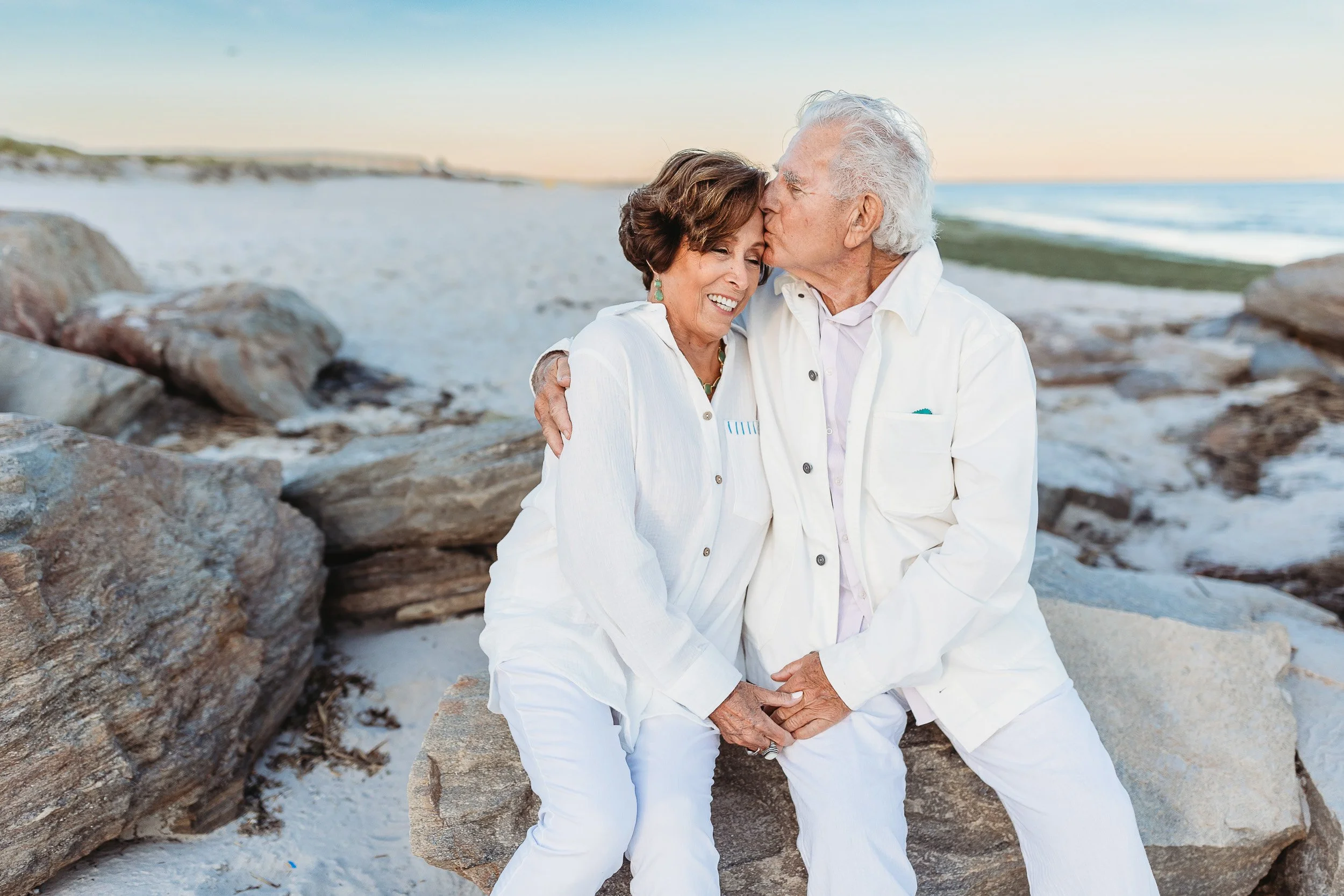 grandpa kissing grandma on the forehand while seated on a jetty at Washington oaks beach 