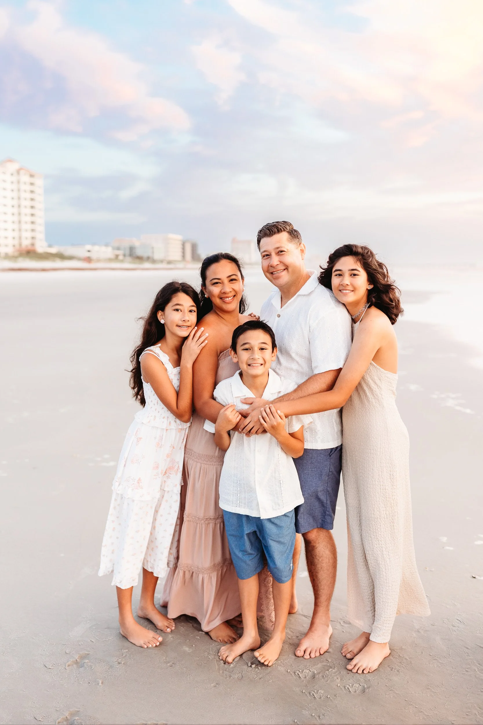 family embraced on the beach at sunrise and they are smiling with the pastel sky behind them