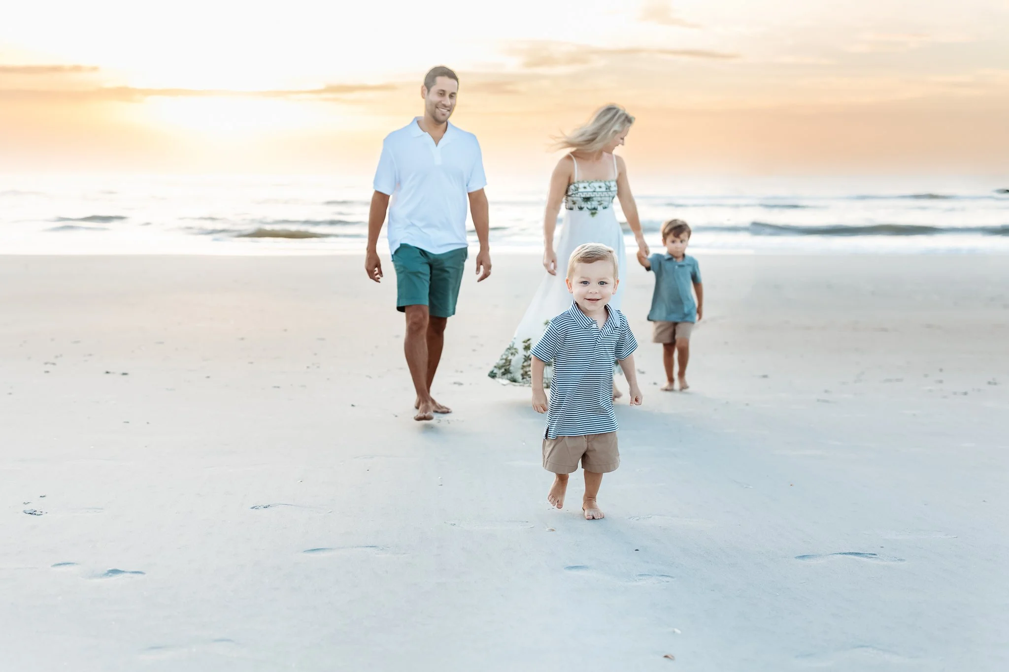 family walking along the beach at sunrise for family photos during a vacation and a toddler baby boy is leading the way