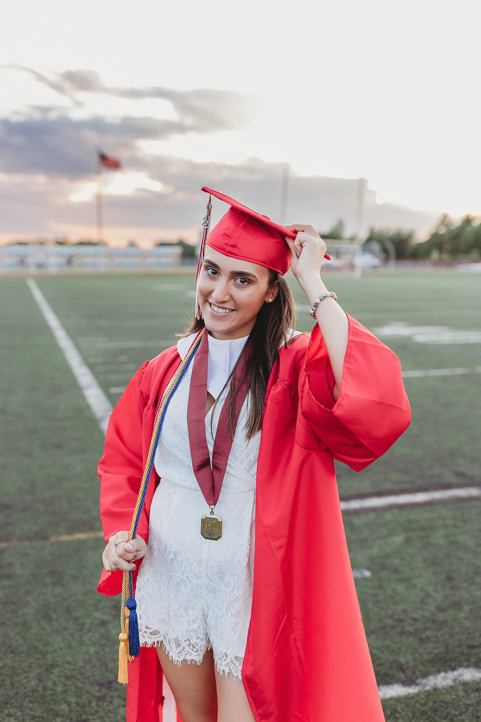 girl in white lace romper on a high school football field wearing red cap and gown for st augustine senior photos