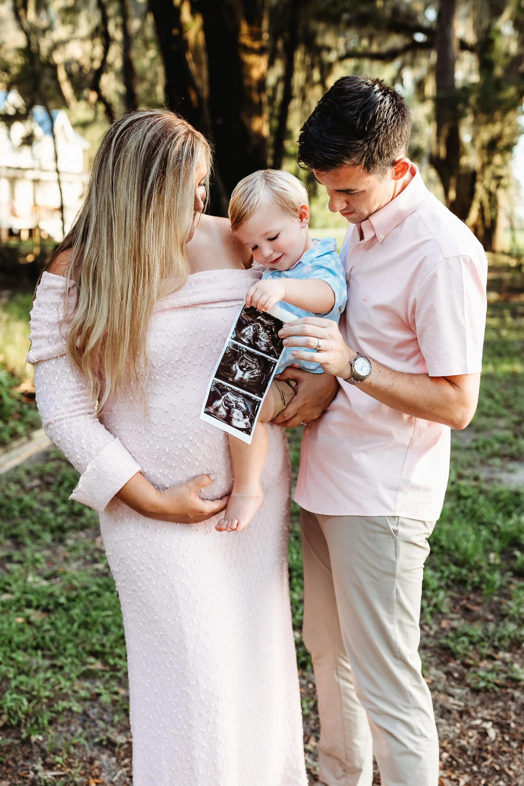 mom holding belly and dad holding toddler while toddler looks at sonogram and laughs