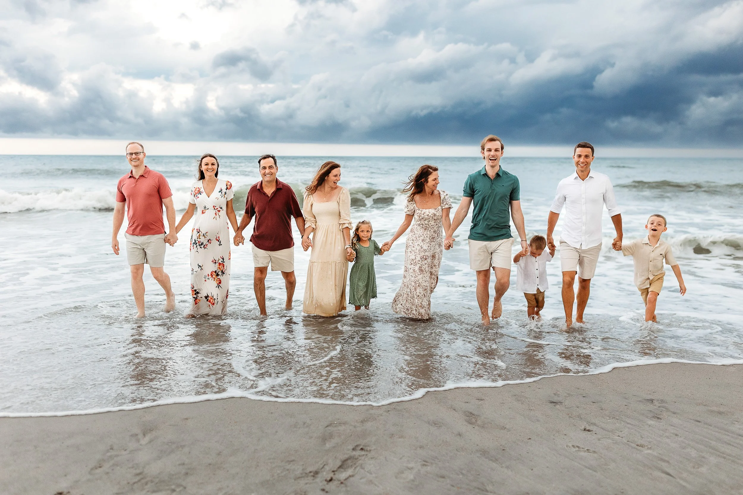 Family walking hand in hand in jewel tones out of the ocean in Villano Beach.  There are grey dar clouds above them and they are laughing 