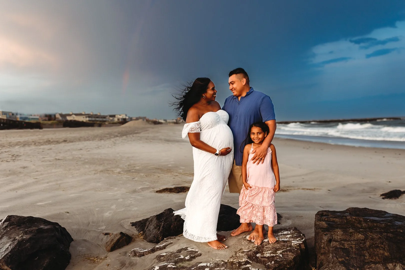 family on a jetty on the beach and mother is pregnant and there is a rainbow behind them