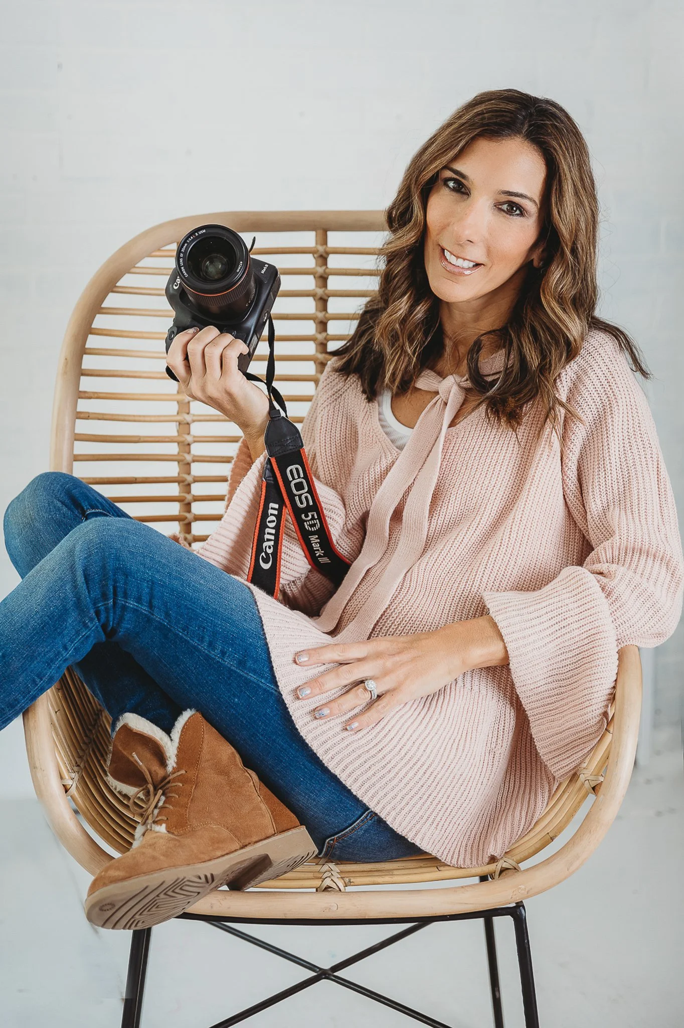A woman sitting in a rattan chair holding a Canon EOS 5D Mark II camera, wearing a pink sweater, blue jeans, and tan boots, smiling at the camera.