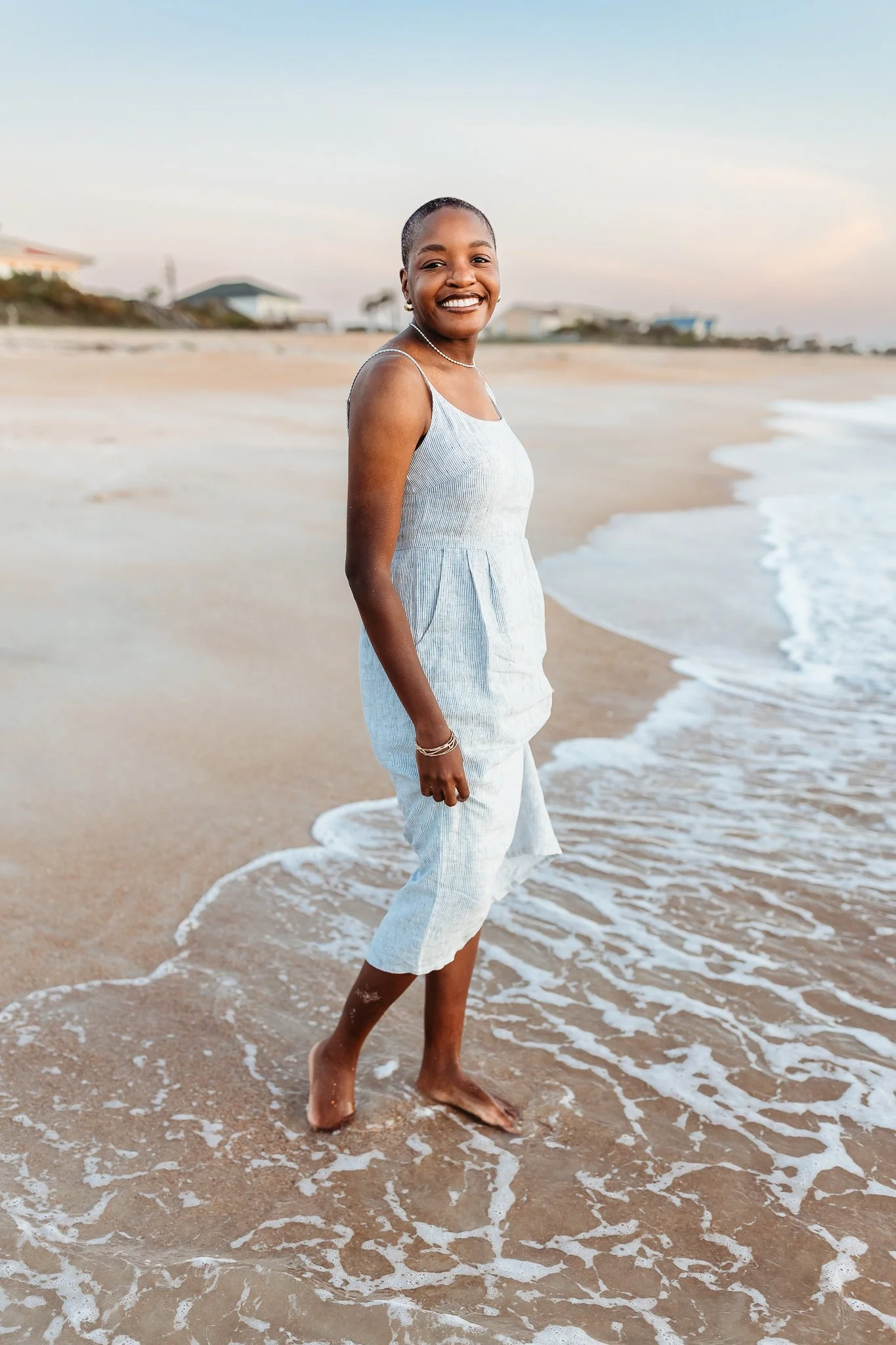 adult daughter in striped linen dress with soft sunrise sky behind her holding dress while standing in the ocean while on vacation in Ponte Vedra Beach