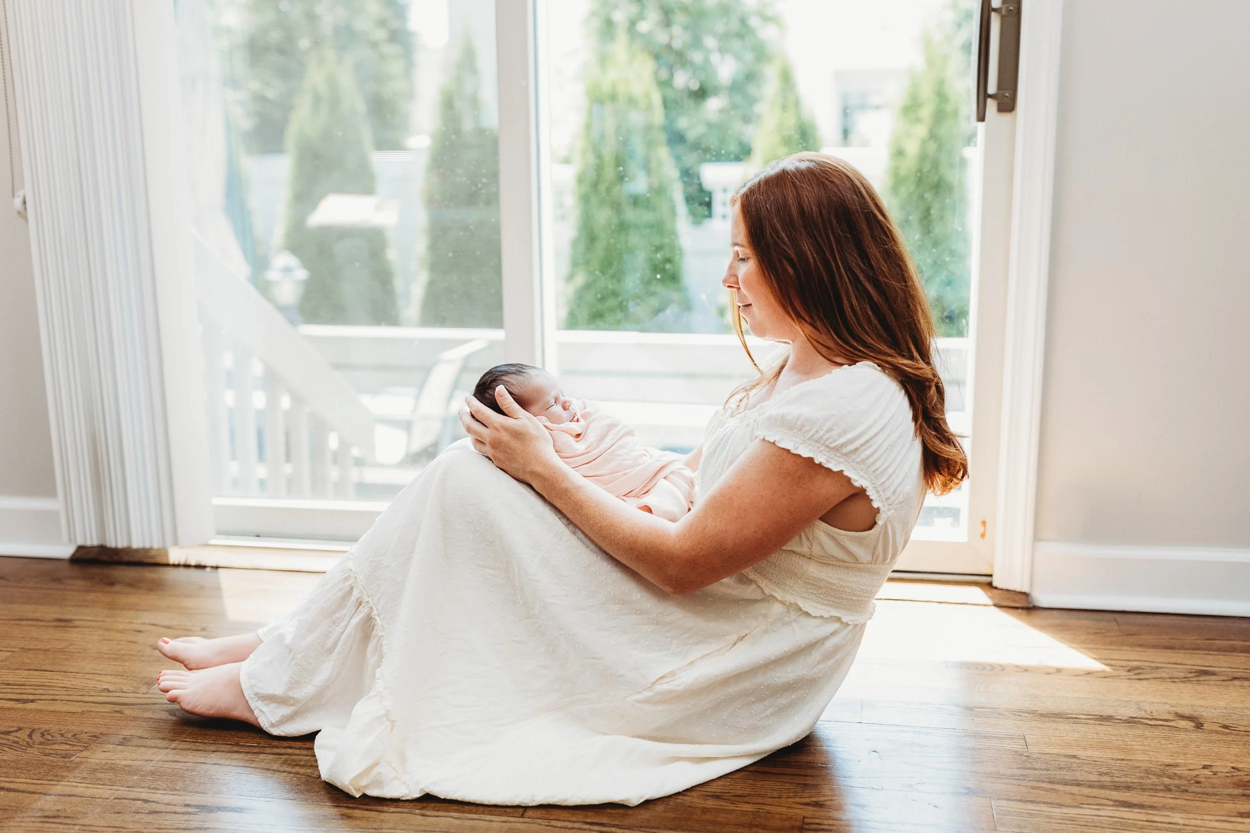 mother in a cream Swiss dot dress holding her newborn swaddled baby on her lap while seated on her floor for her in home newborn photos