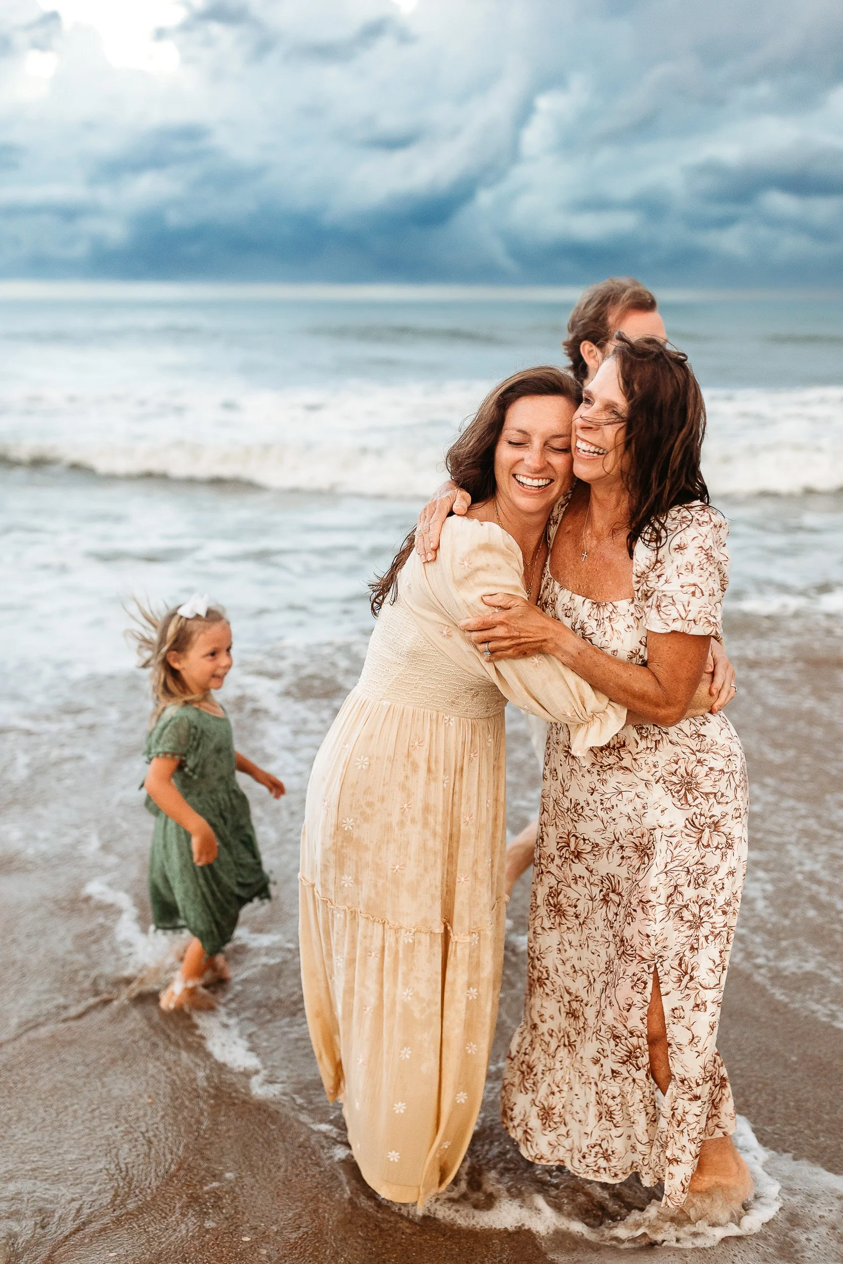 candid moment of a mother embracing her adult daughter in the ocean at Jacksonville beach.  they are both laughing and the granddaughter is frolicking in the waves behind them 