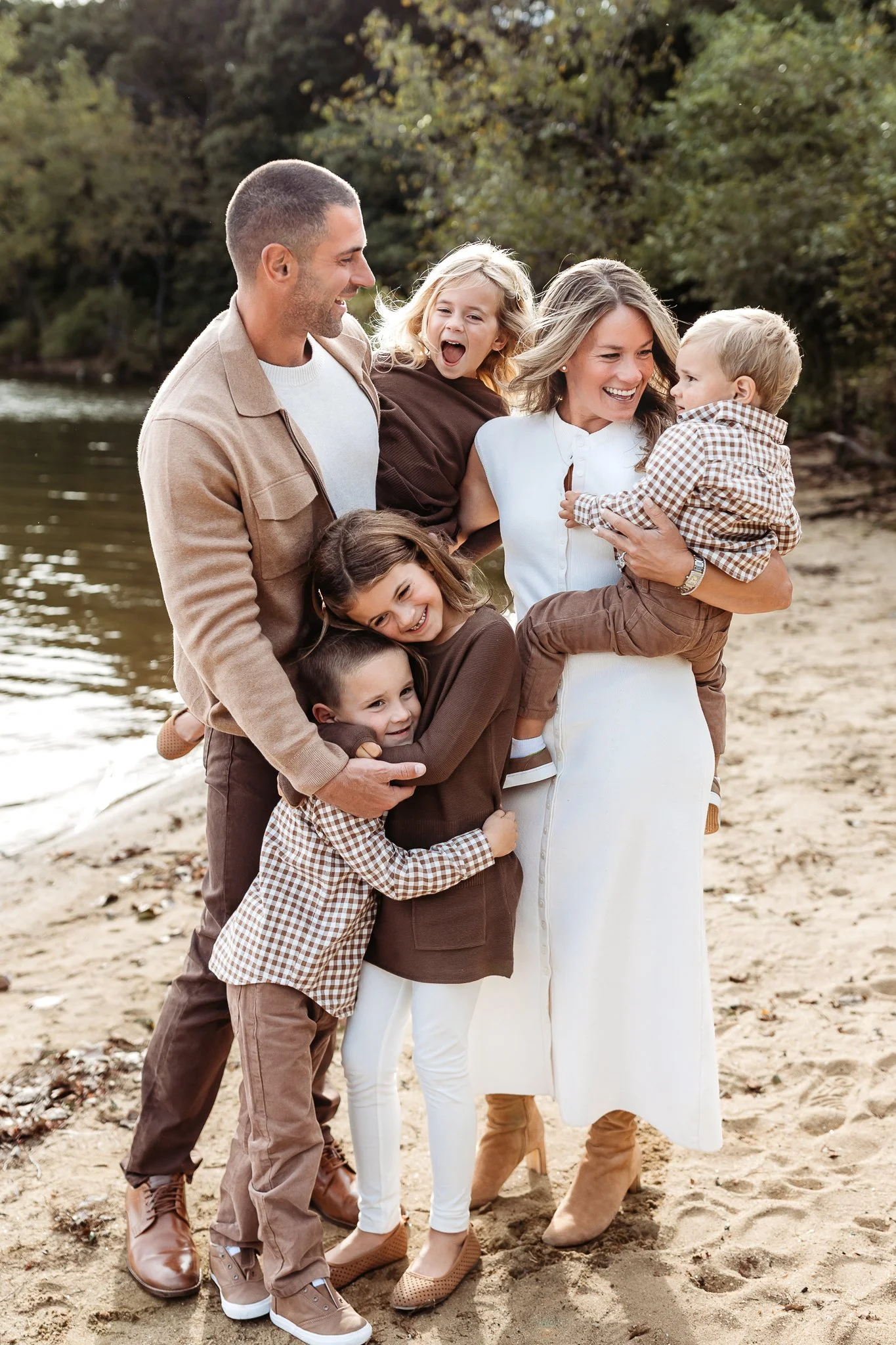 Parents embracing their four children beside the water, wearing brown, camel, and cream.