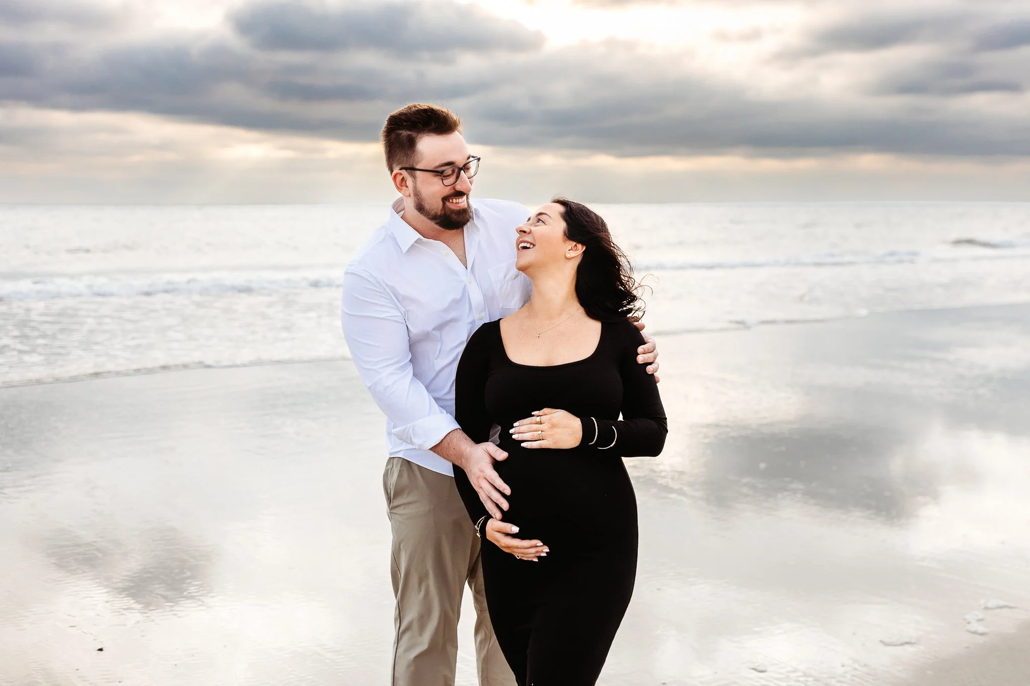 Pregnant mother leaning into her partner with their hands resting together on her belly.