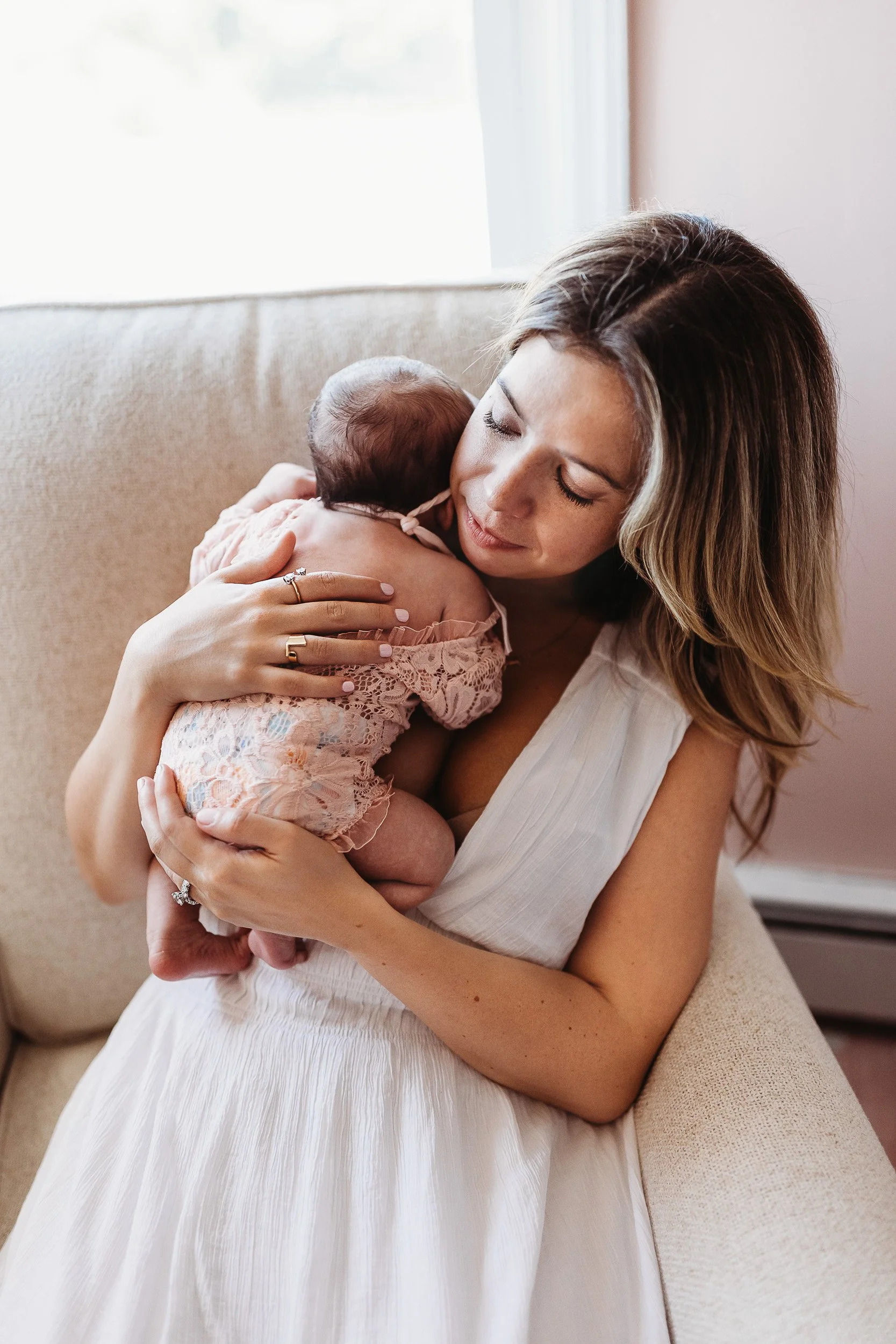 mother state din a glider in a nursery for her in home newborn photo session with a baby girl nuzzled up on her shoulder and baby is sleeping while swaddled and there is a window with lovely light behind them
