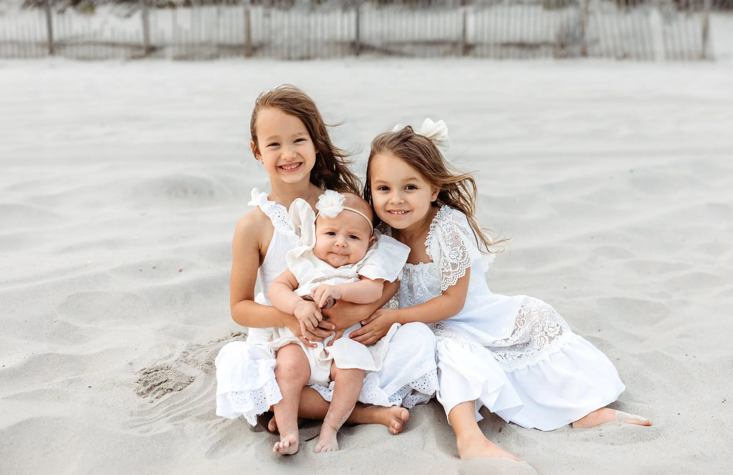toddler siblings dressed in white seated on the beach for family photos and they are holding their new baby sister and smiling