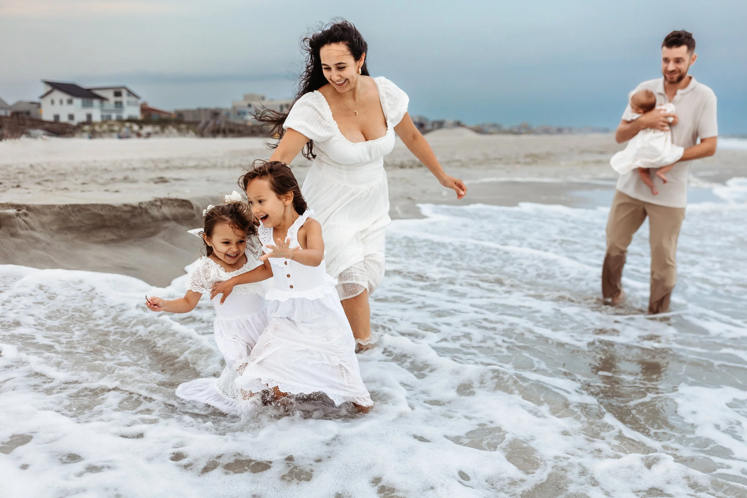 mom playing in the waves with sibling girls and dad inholding the baby laughing