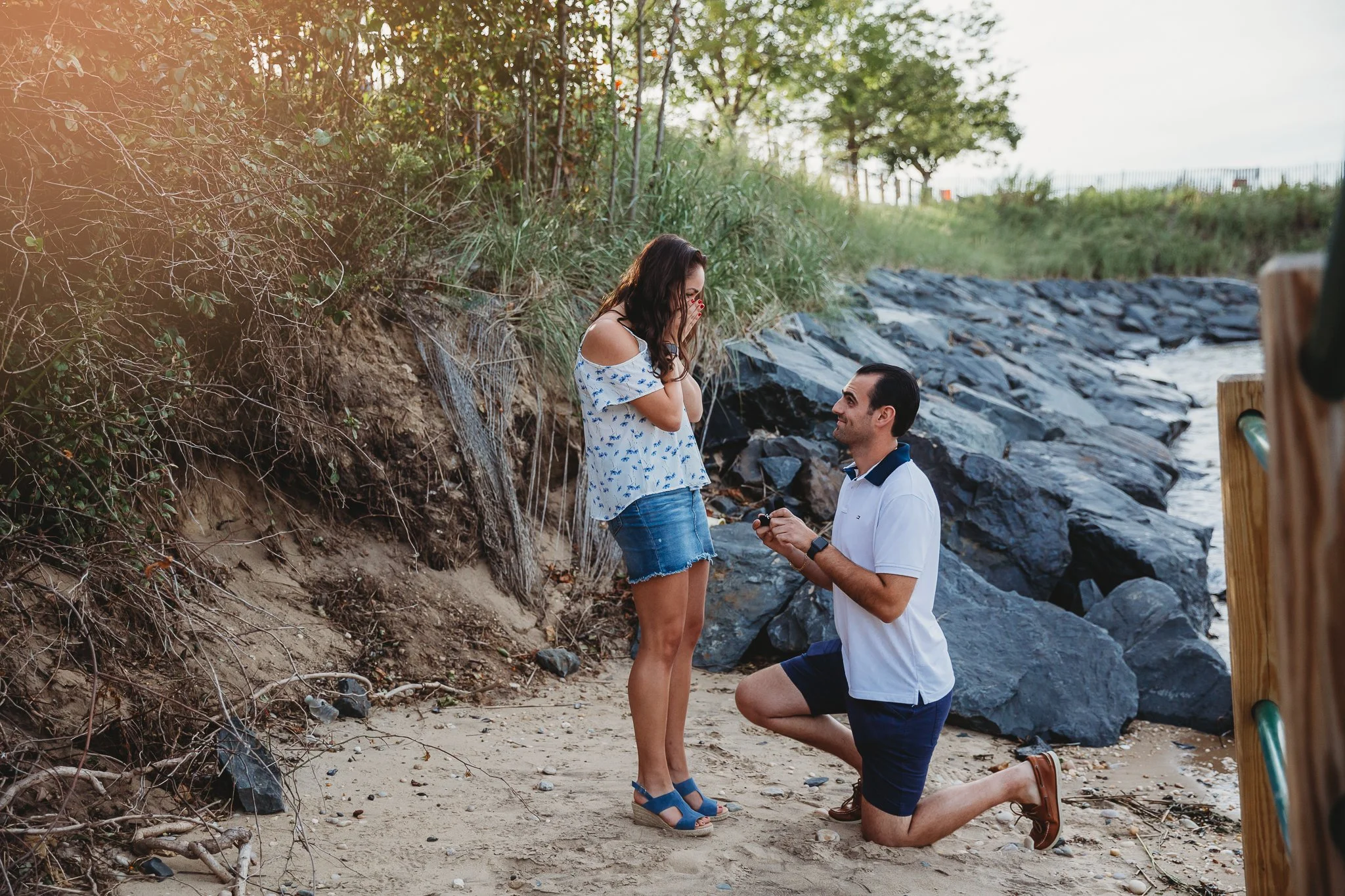A man proposing marriage to a woman on a beach at sunset, with the woman appearing emotional and the man holding a ring.