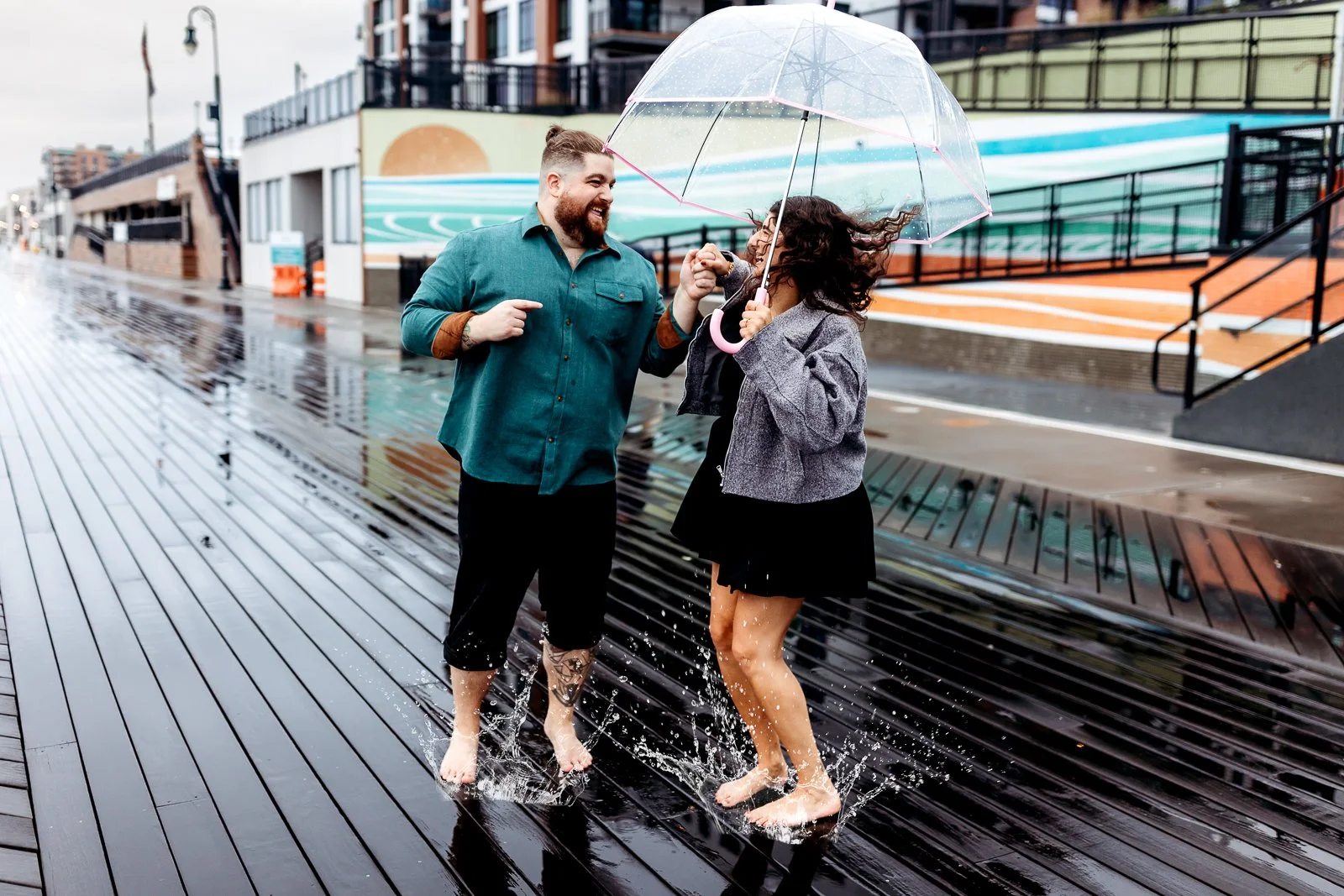 A man and woman are smiling and playing with water on a wet outdoor boardwalk, holding a transparent umbrella during a rainy day.