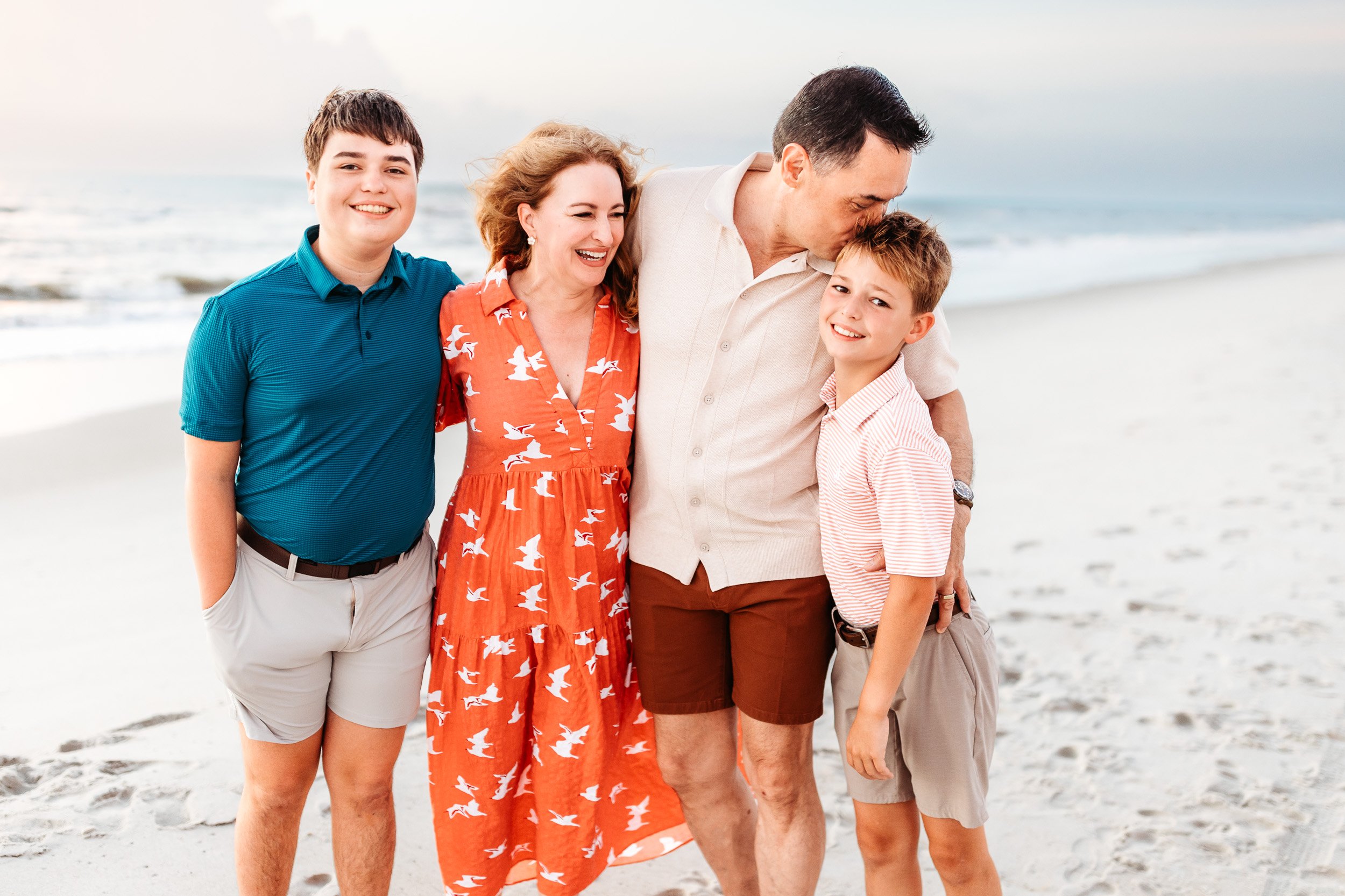 dad kissing son and mom is laughing they are at the beach at sunrise