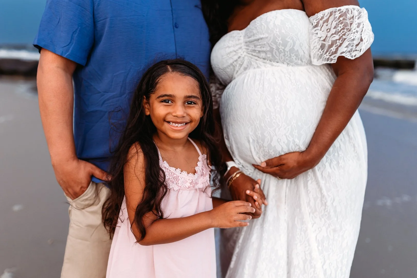 close up of a little girl in front of a mom's pregnant belly and she is smiling mom is holding her belly and they are on Jacksonville Beach