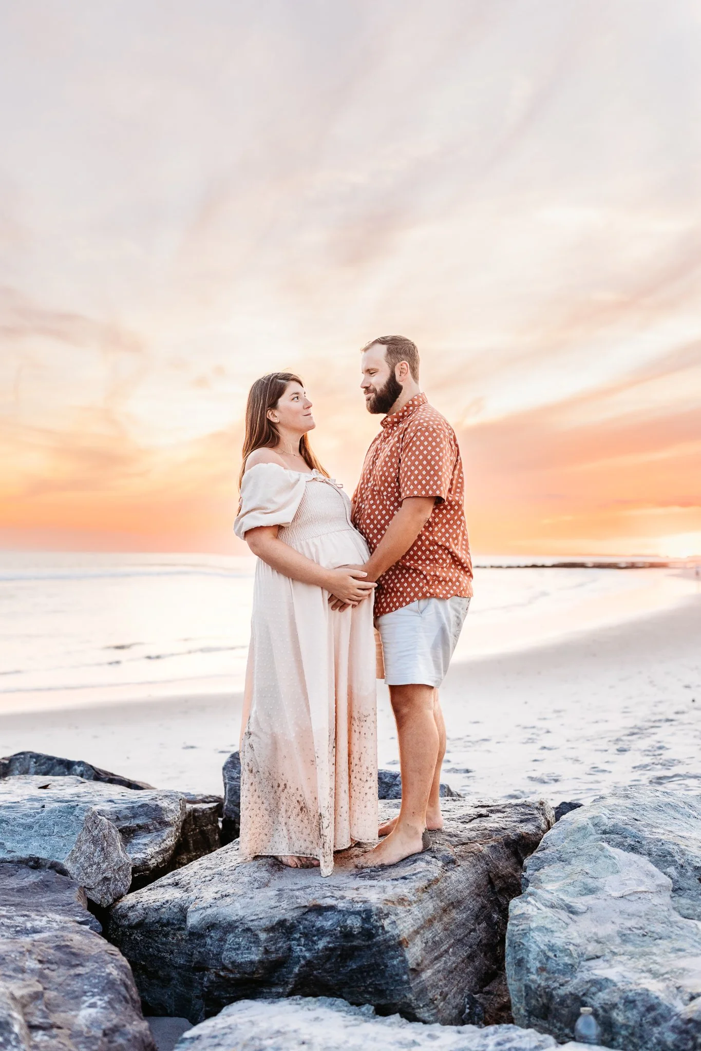 couple standing on a jetty for Jacksonville maternity pictures and woman is in the free people oasis dress and man looking at woman with sunset over the ocean behind them