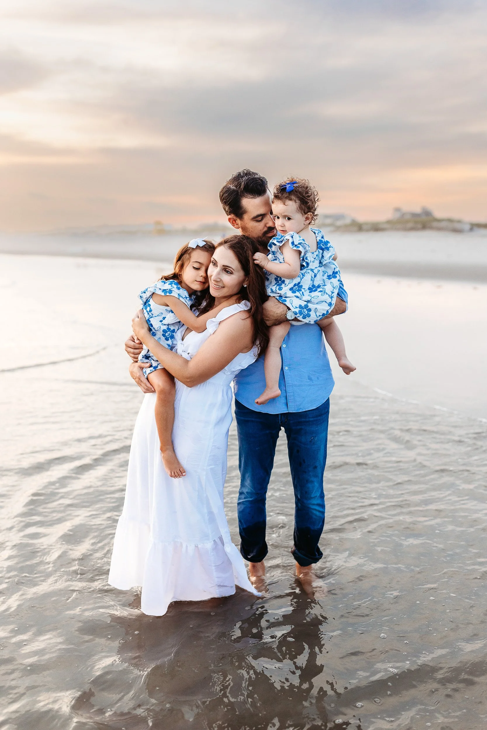 connected moment between a family during an Atlantic Beach photo session.  the parents are holding their toddlers and kissing them and the sunset is a pretty orange color.  they are wearing blue and white outfits 
