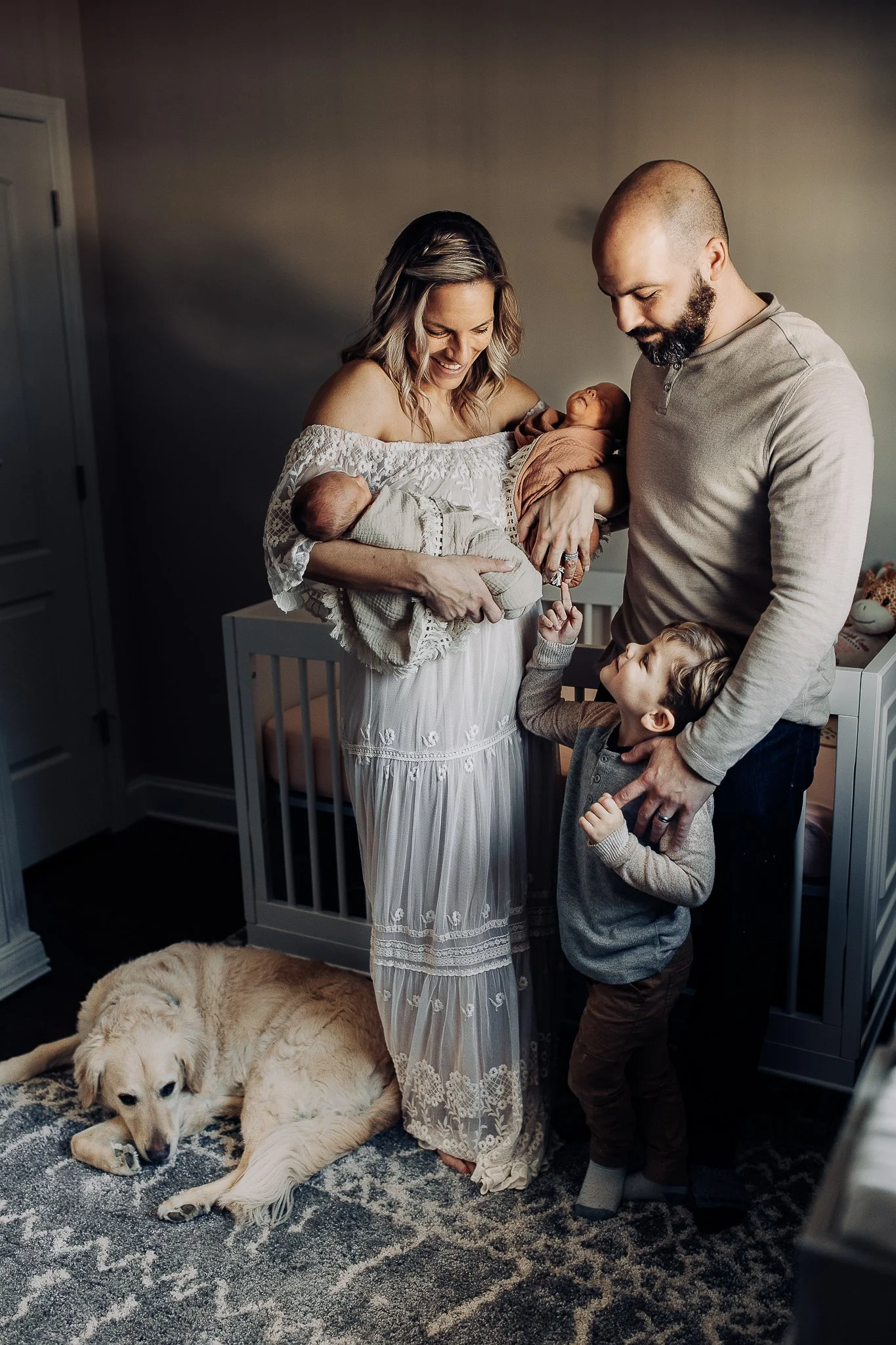 family in a nursery and mom is in a lace gown holding twins during in. home Jacksonville newborn pictures and a golden retriever is on the floor