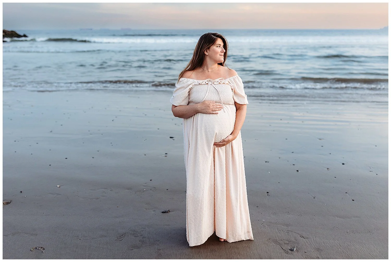 pregnant woman holding belly ocean behind her looking at horizon