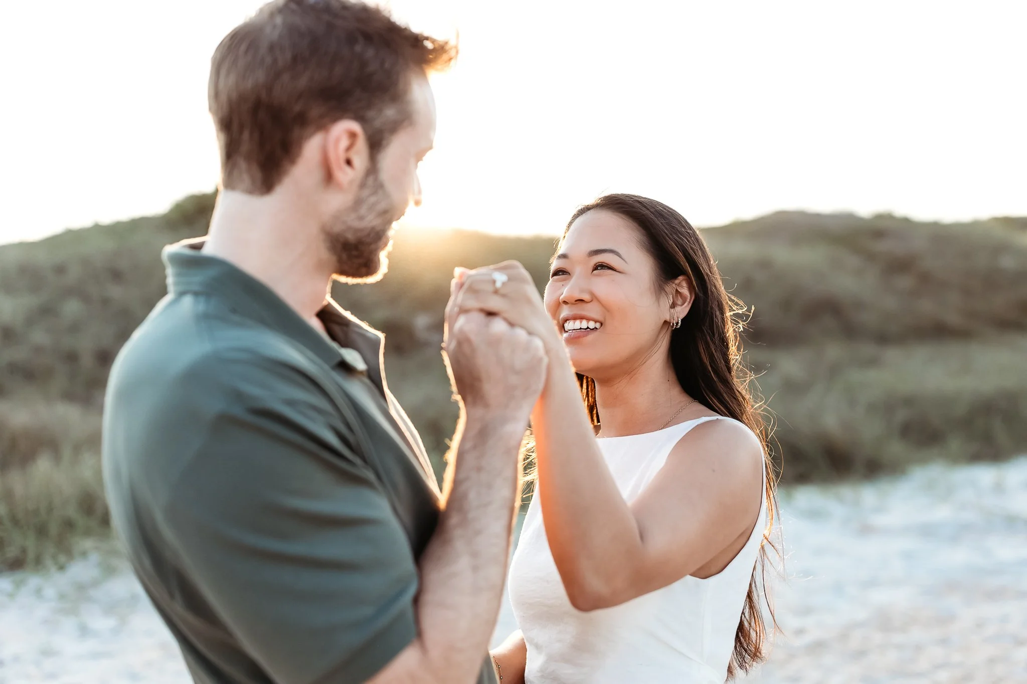 man holding hand of woman with engagement ring on at sunset in front of a dune at guana preserve and she is looking at him and smiling