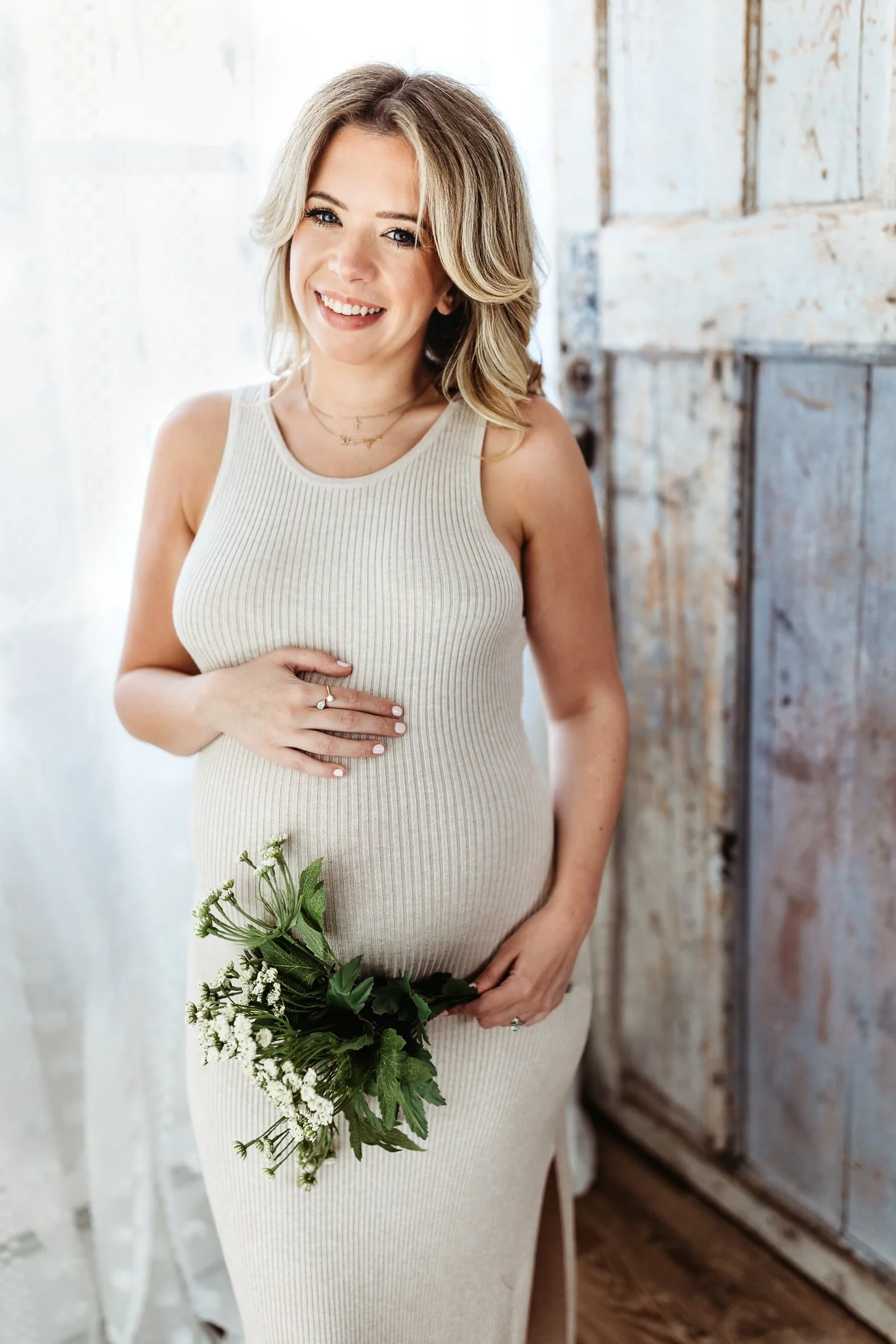 woman who is pregnant in a knit dress leaning against a wood door holding a. bouquet of flowers
